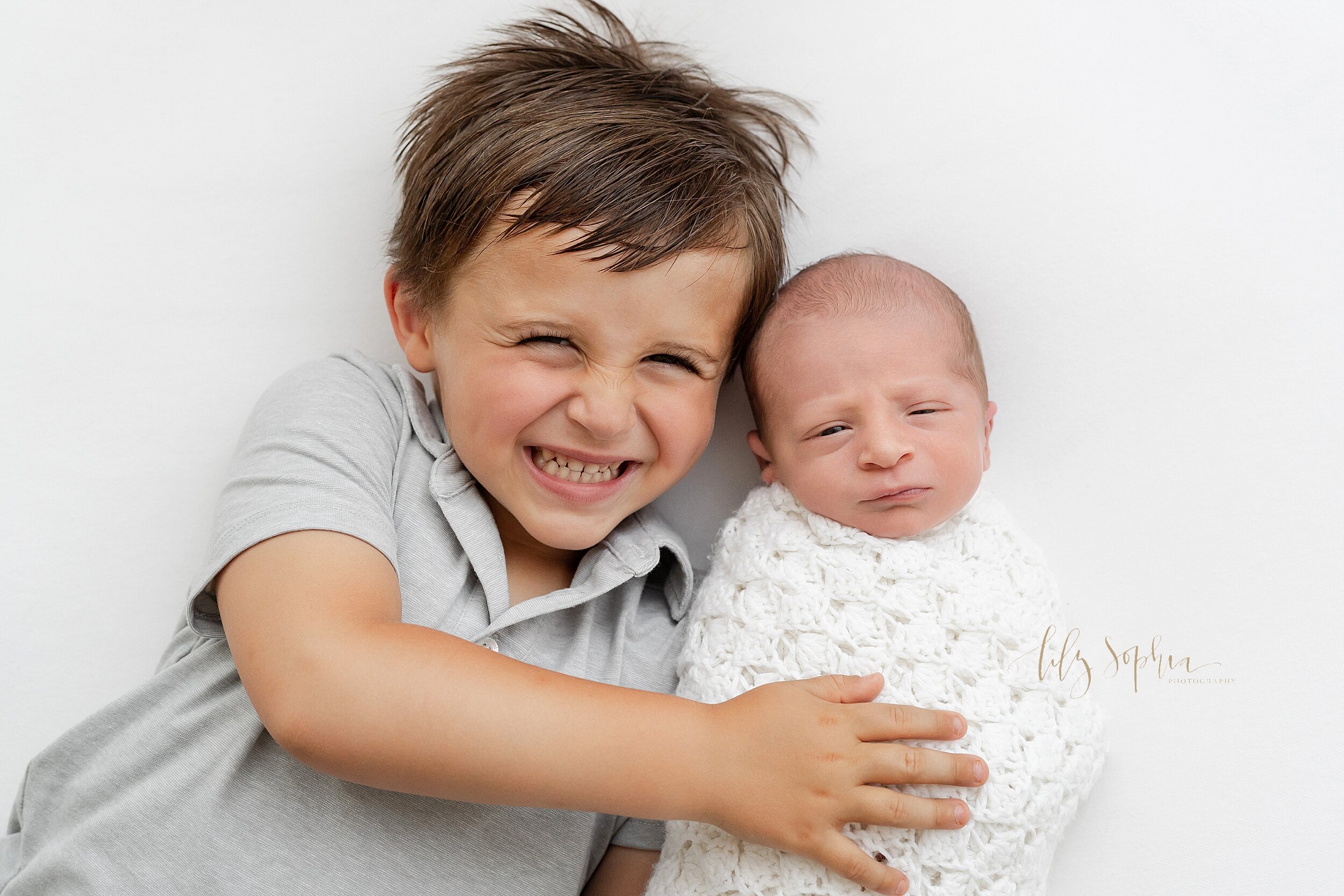  Family photo with a smiling young boy lying next to his awake newborn brother as they lie on their backs in a studio near the Brookhaven area of Atlanta, Georgia in natural light. 
