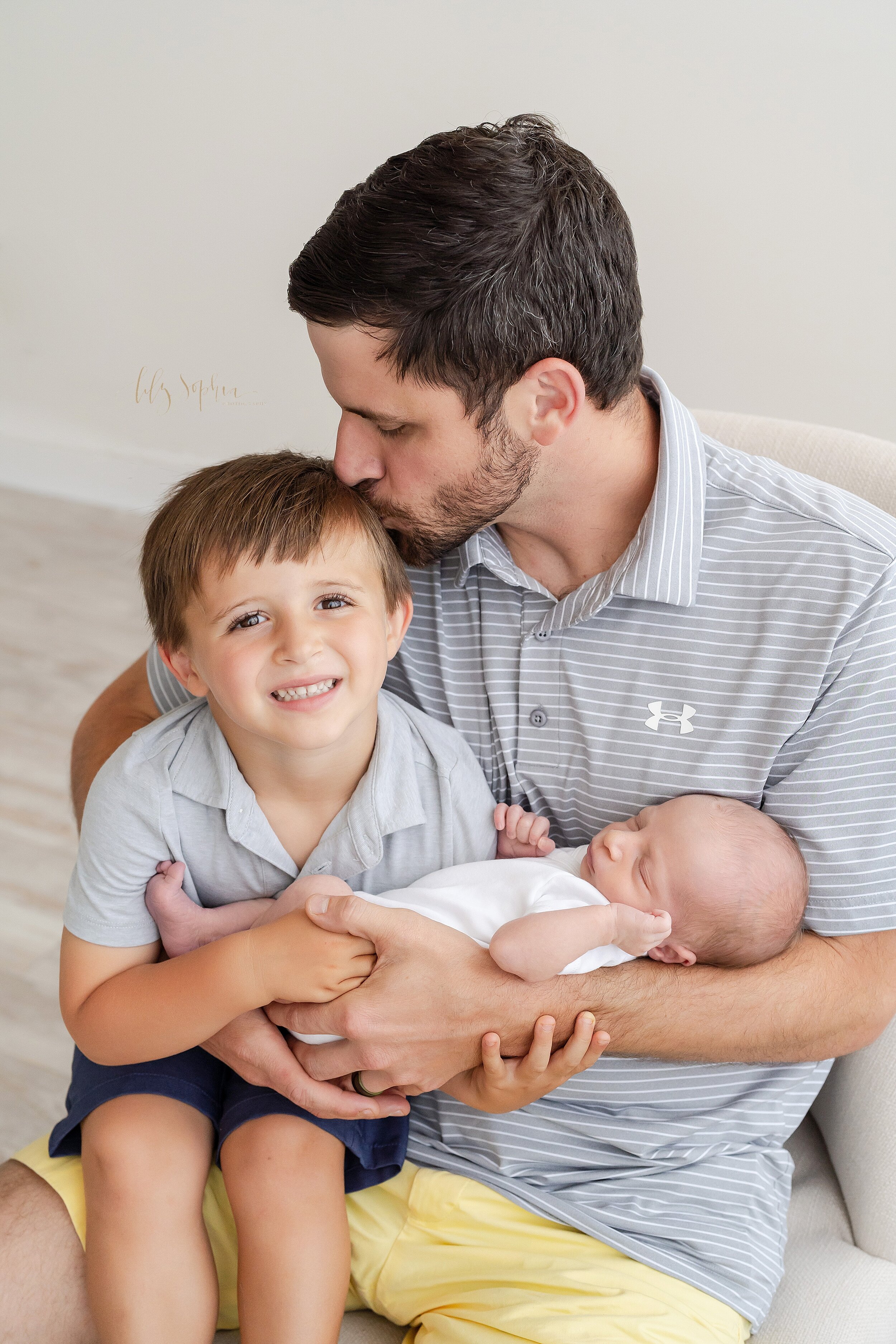  Family photo of a father holding his young son as the son helps to hold his newborn brother with Dad kissing the top of his young son’s head as they sit in a natural light studio in the Ansley Park area of Atlanta. 
