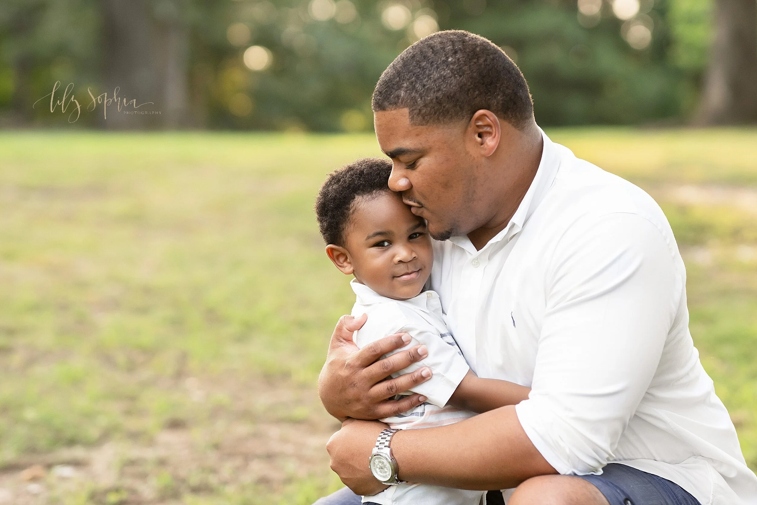 Family photo shoot in a park in Atlanta, Georgia of an African-American father as he holds his toddler son tightly as he crouches to embrace him at sunset in an Atlanta park.