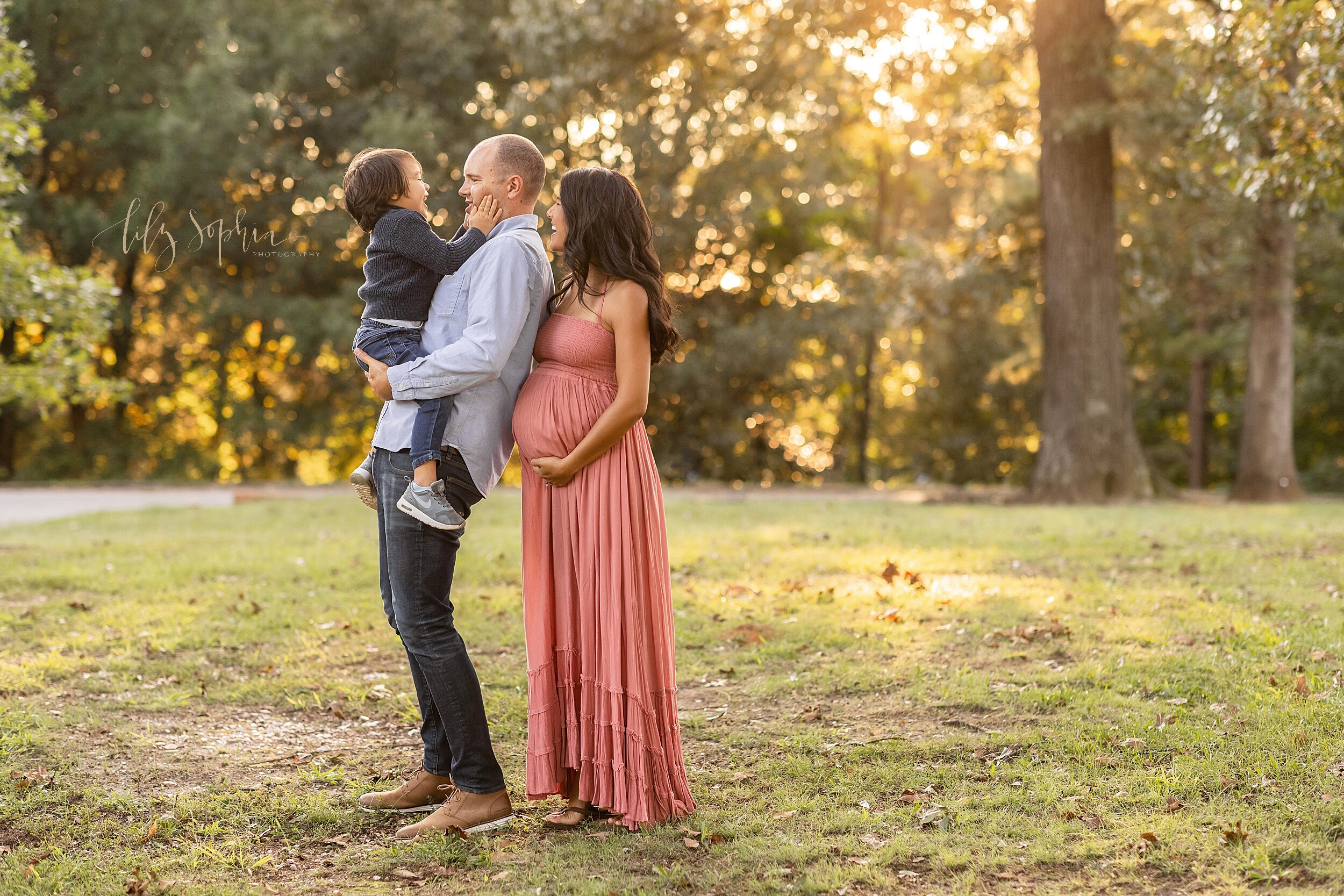  Maternity photo session of a father holding his son who is facing him and putting his hands on his cheeks while his expectant wife stands holding the bottom of her belly behind him and peeks over his left shoulder as the sun is setting in a park nea