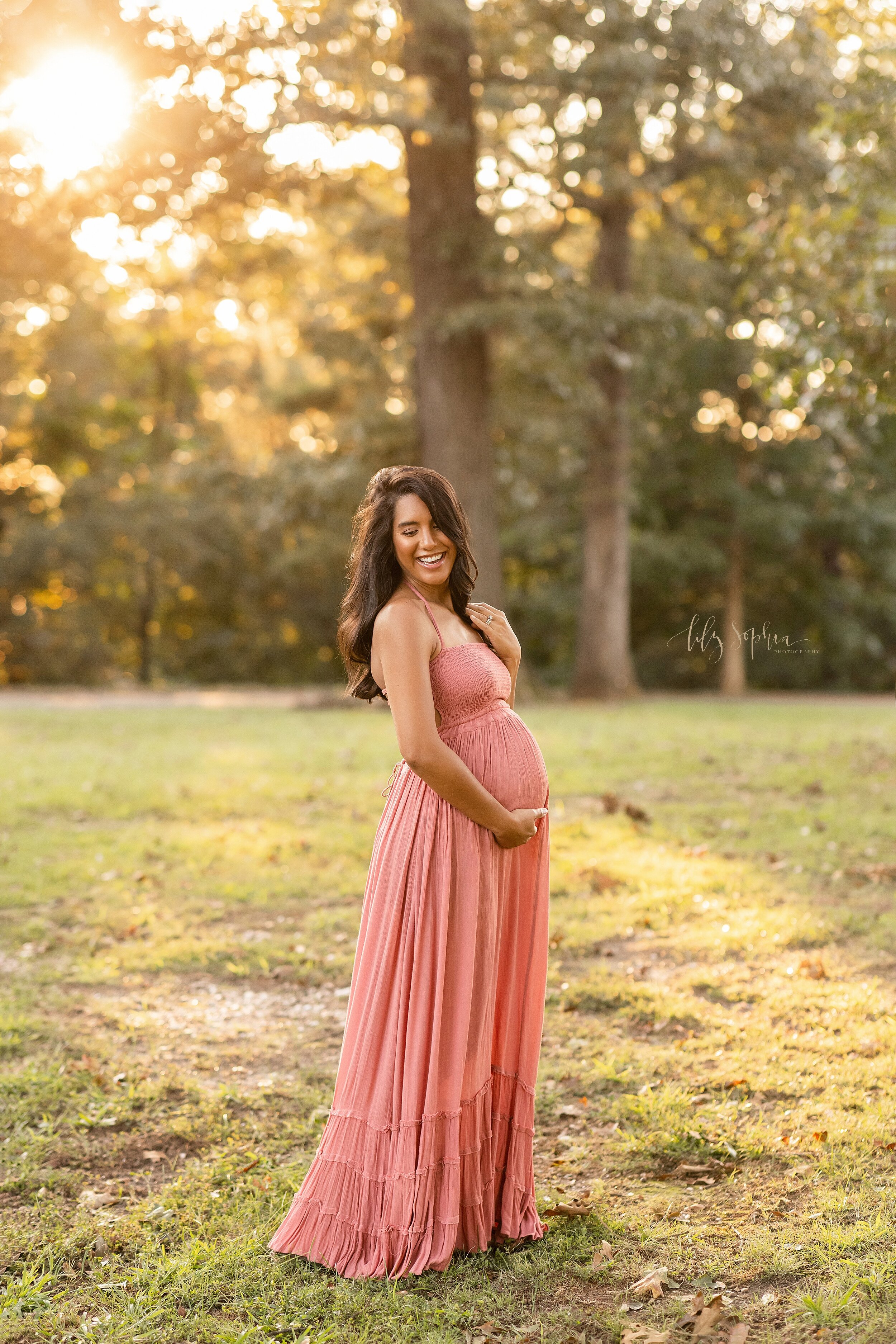  Maternity portrait of a brunette pregnant woman as she stands at sunset in a park and frames her belly with her hands as she looks over her right shoulder near Atlanta, Georgia. 
