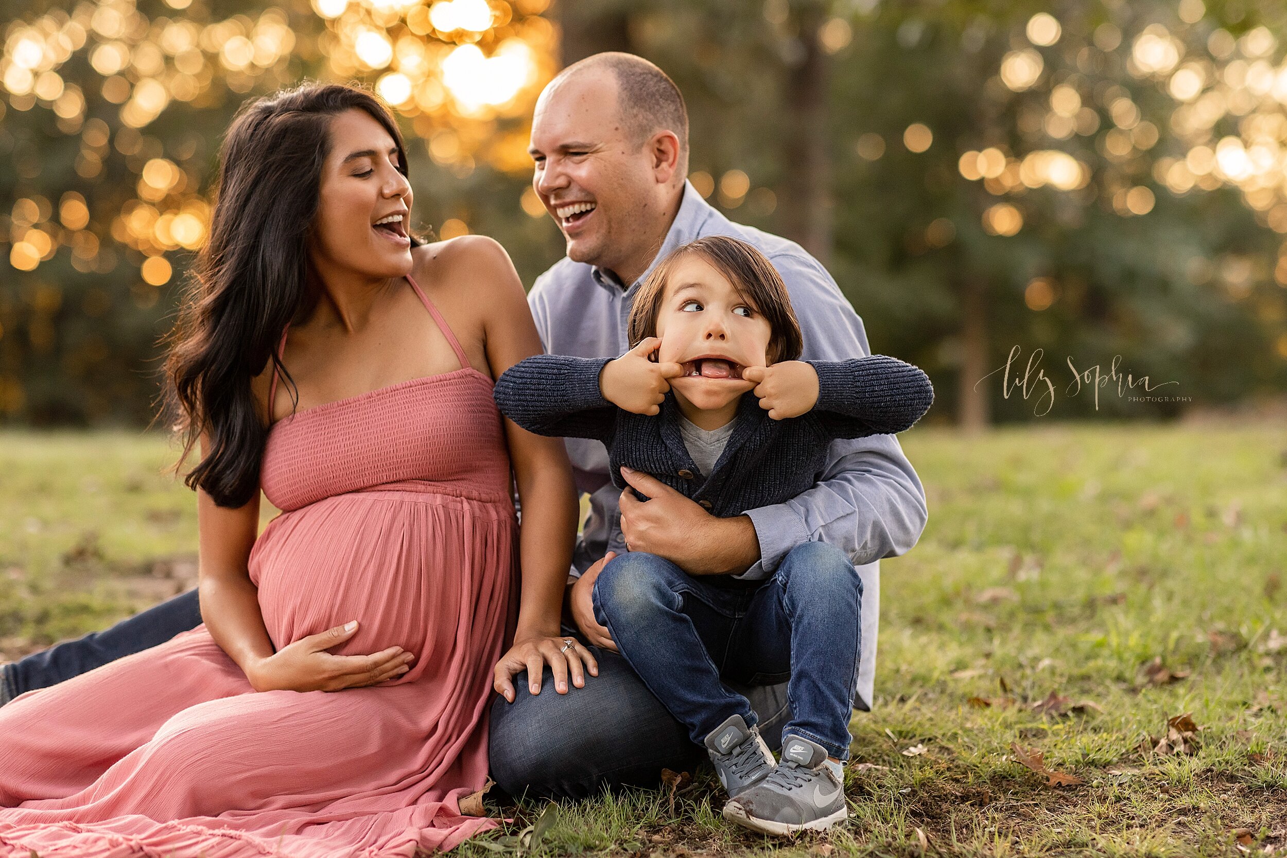  Maternity photo of a mom sitting on the grass in a park next to her husband who is holding their young son on his lap as his son makes a funny face taken at sunset in an Atlanta park. 