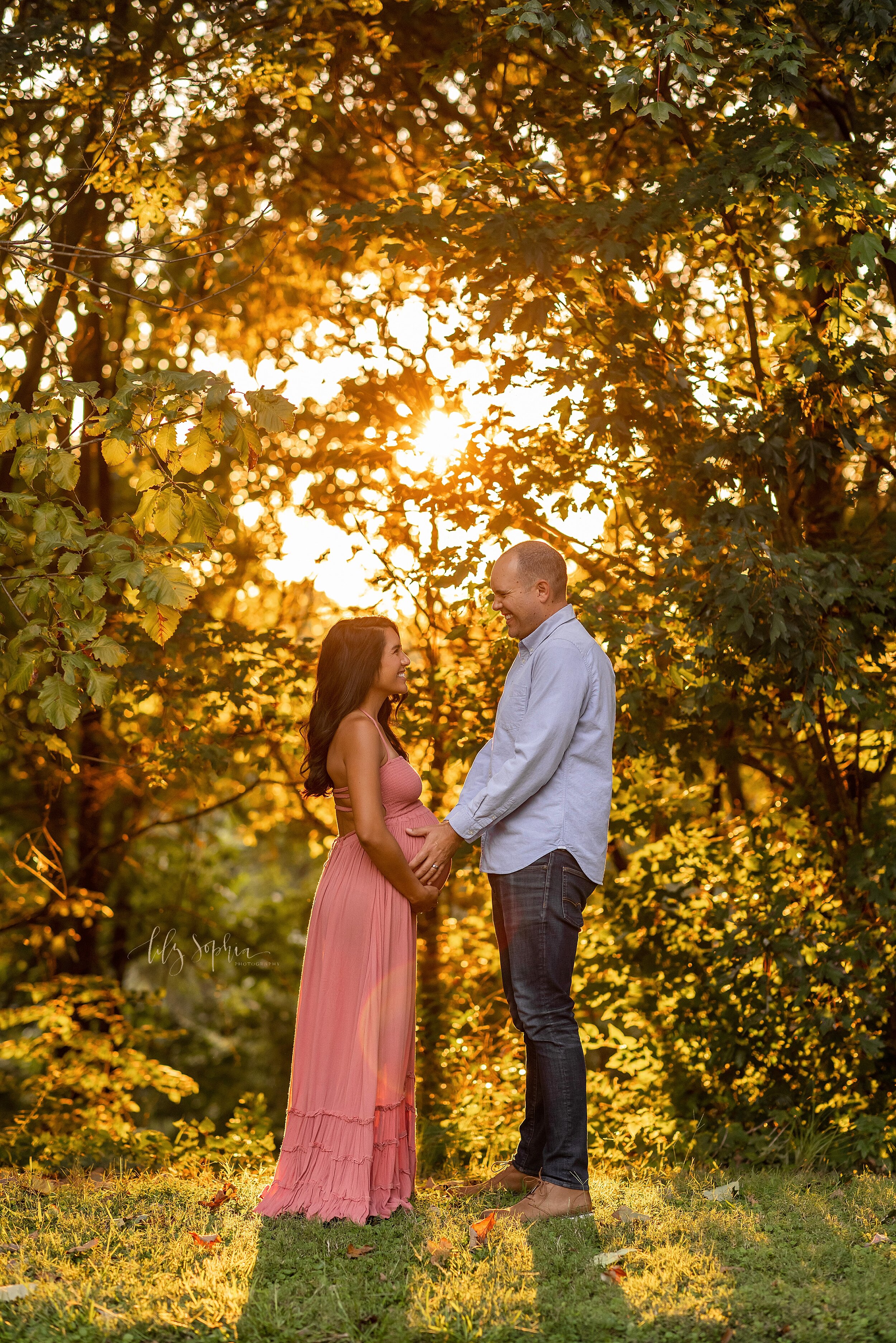  Maternity photo session in an Atlanta park of a husband and wife facing one another as they lovingly make eye contact and both of them hold their child in utero with the sun setting. 