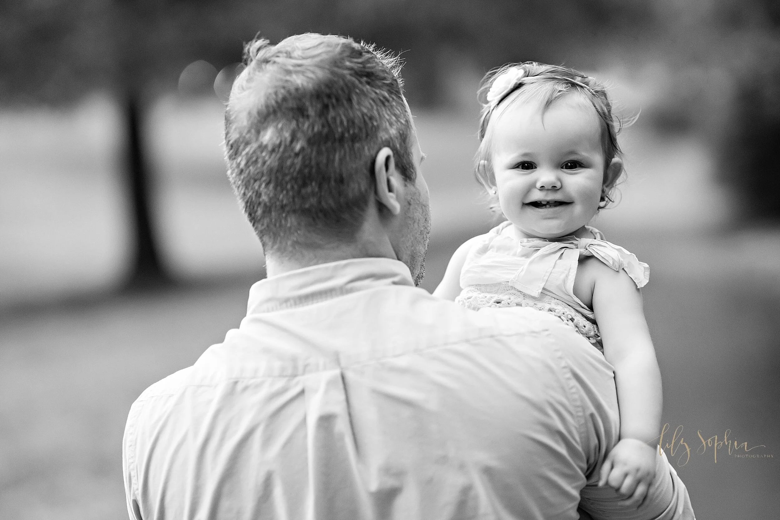 Black and white image of a father holding his one year old daughter in his arms as she smiles while looking over his shoulder as they walk in a park in the Vinings area of Atlanta.