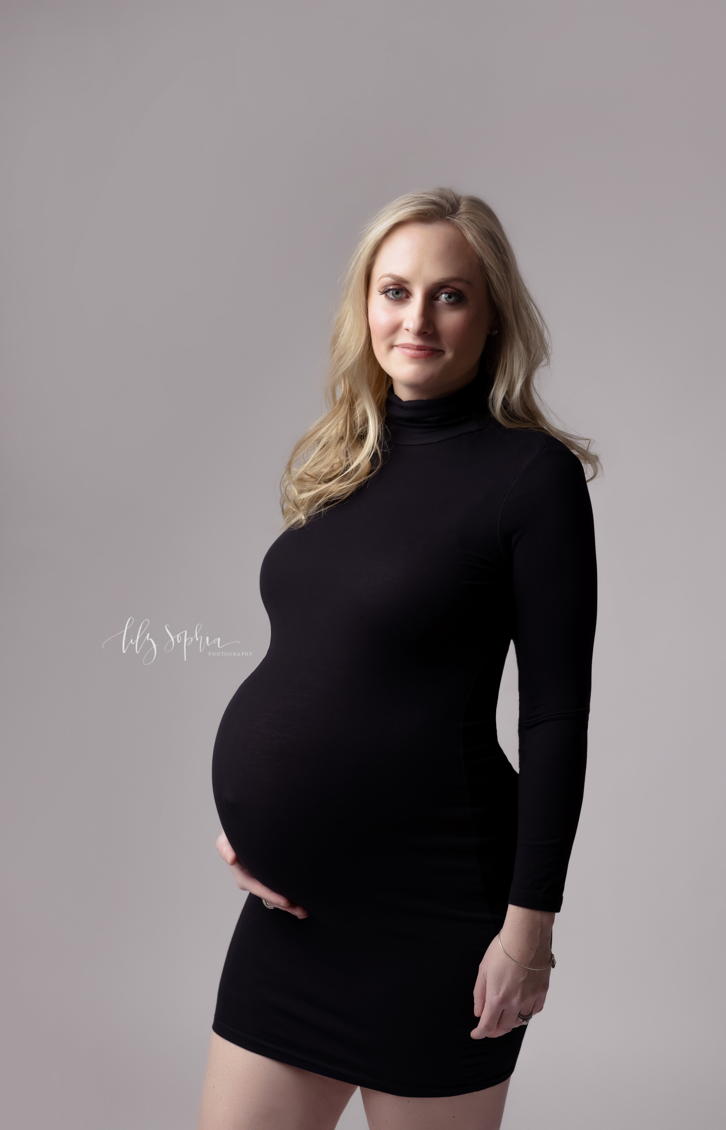  Color  editorial maternity fine art image of beautiful smiling pregnant woman with long blonde hair dressed in black turtleneck bodycon dress on a gray background in the Atlanta studio of Lily Sophia Photography.  