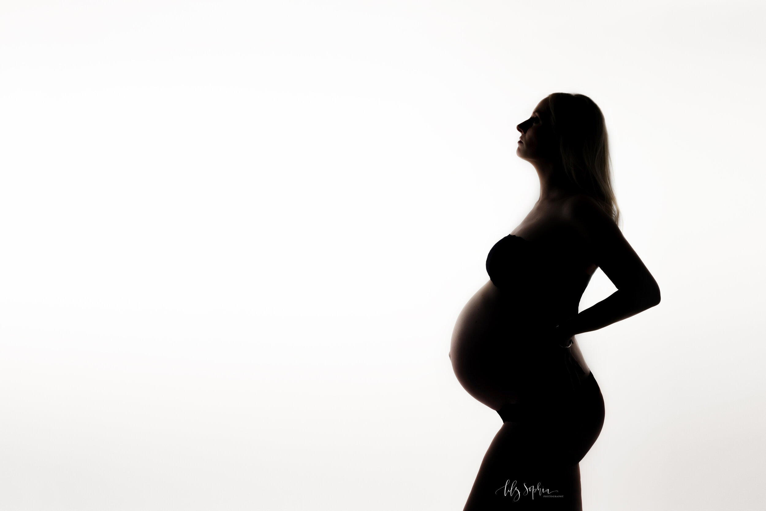  Silhouette editorial maternity fine art image of beautiful pregnant woman with long blonde hair dressed in black lace bra and underwear against a white background in the Atlanta studio of Lily Sophia Photography.  