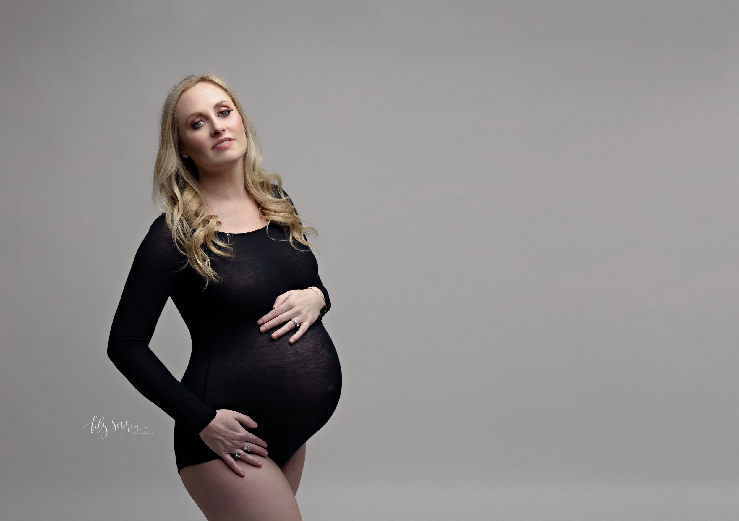  Color  editorial maternity fine art image of powerful 37 weeks pregnant woman with long blonde hair dressed in black bodysuit against  a gray background in the metro Atlanta area studio of Lily Sophia Photography.  