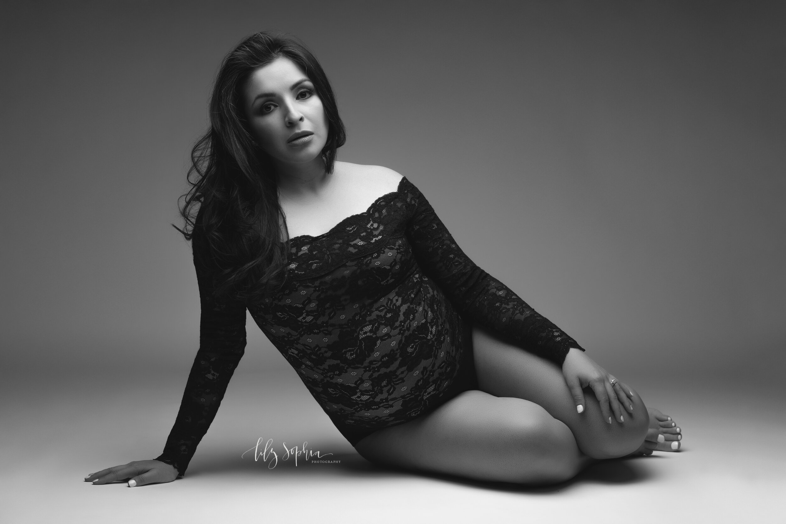  Black and white maternity fine art image of beautiful pregnant Hispanic woman with long dark hair dressed in black lace bodysuit sitting on floor in the Atlanta studio of Lily Sophia Photography.  