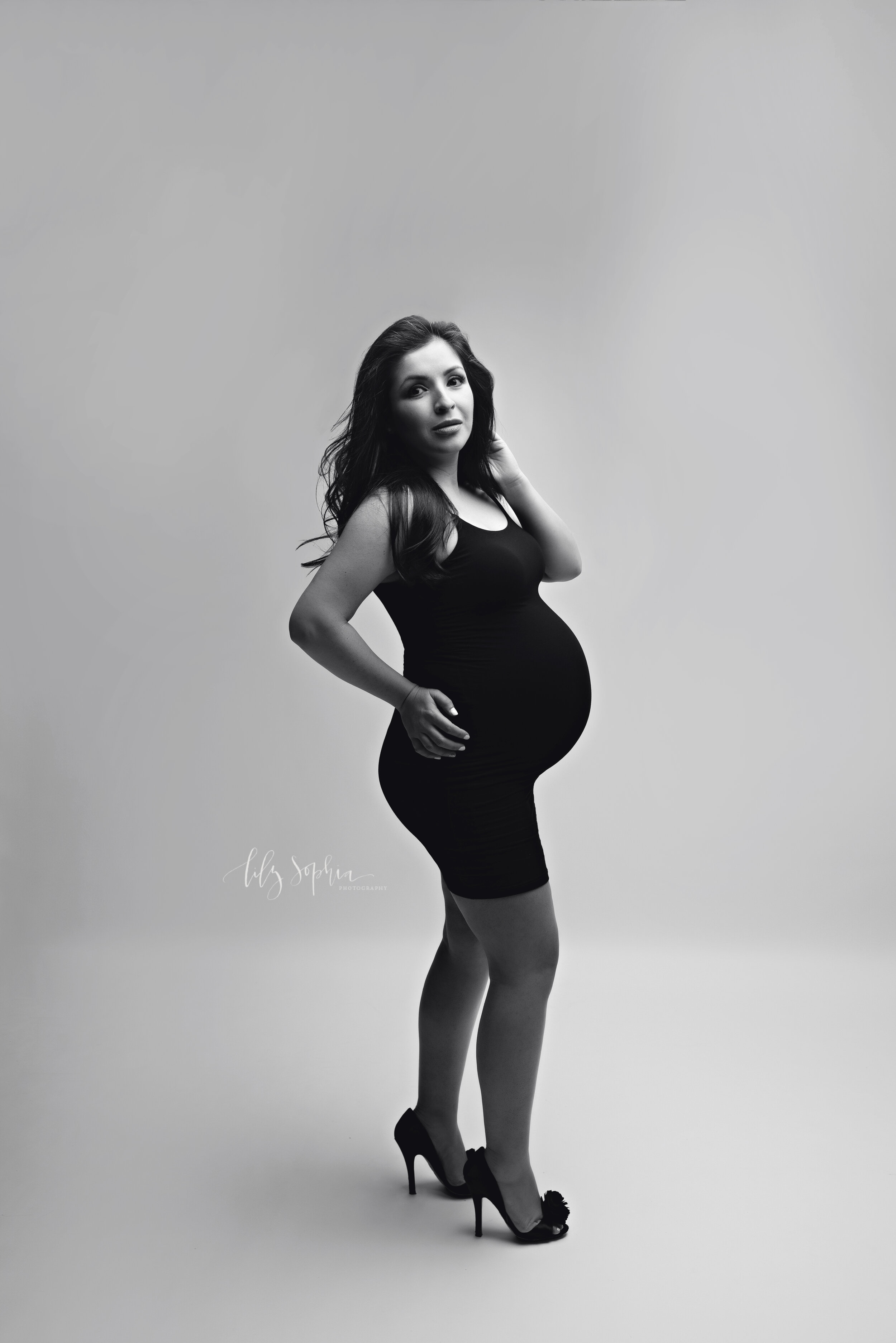  Black and white editorial maternity fine art image of beautiful pregnant Hispanic woman with long dark hair dressed in black bodycon dress and heels on a white background in the Atlanta studio of Lily Sophia Photography.  