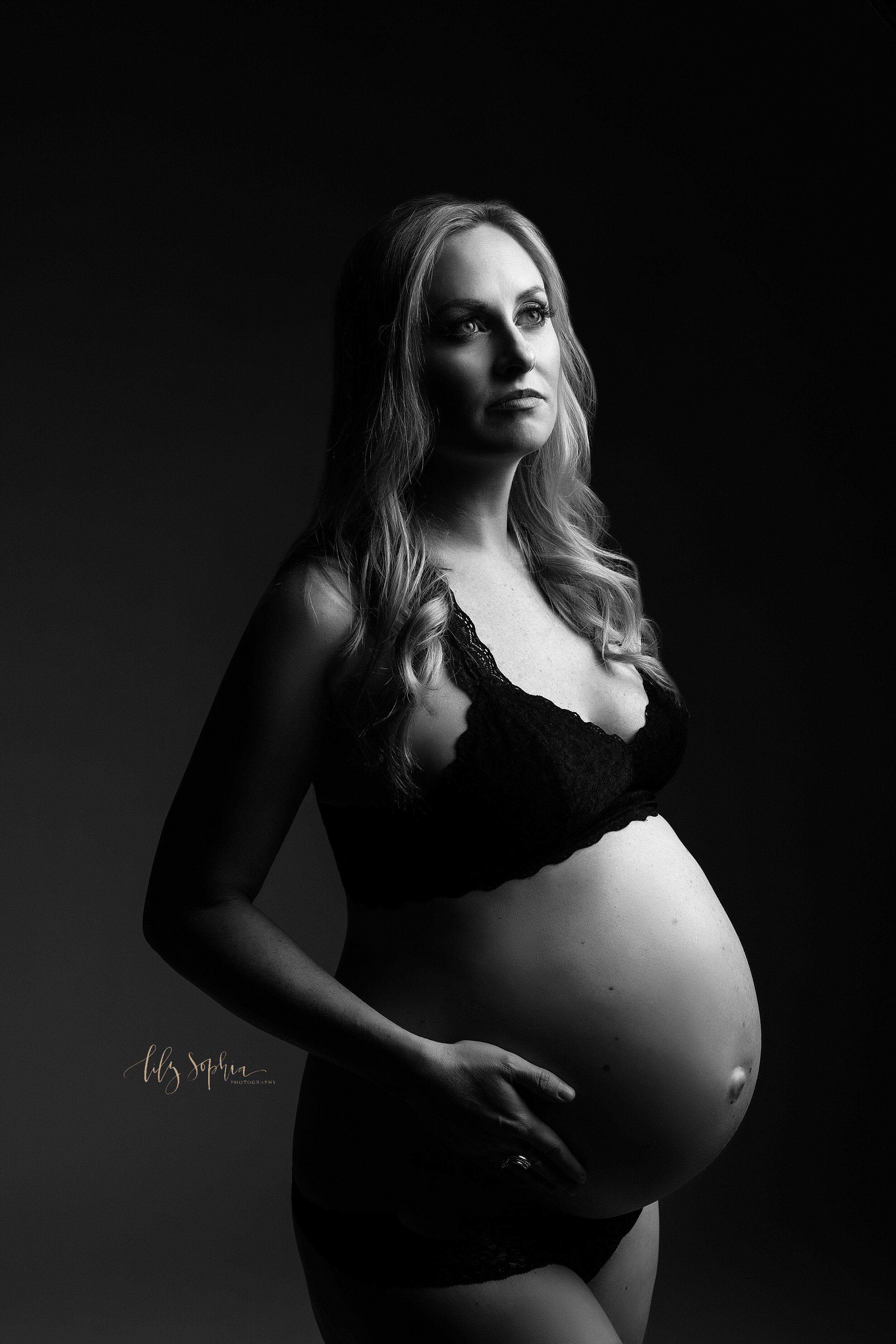  Black and white fine art portrait of blonde pregnant woman in Atlanta studio wearing a black lace bra and panty set with one hand on her belly looking up towards the light. 