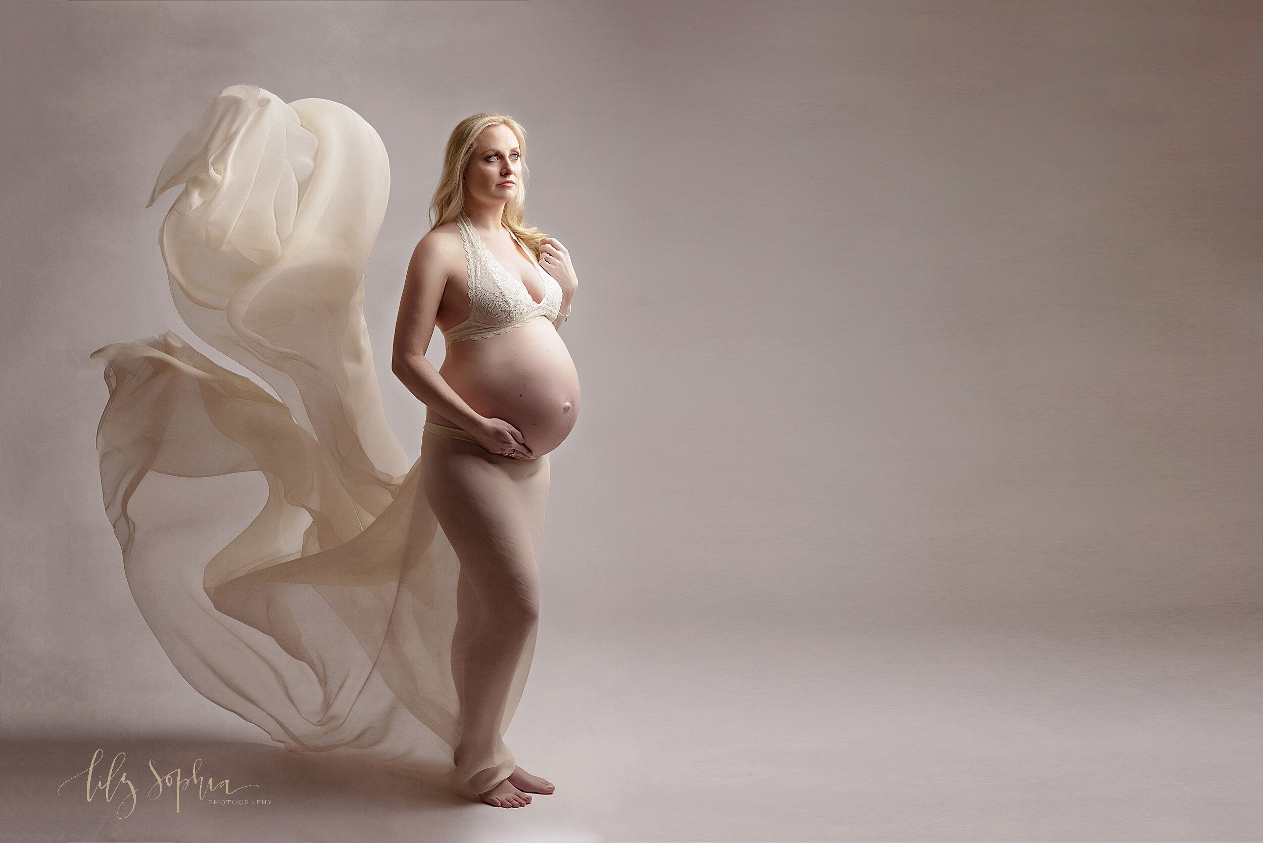  Fine art maternity portrait of blonde pregnant woman in the Atlanta studio of Lily Sophia photography wearing an ivory lace bra with tossed floating chiffon fabric behind her. 