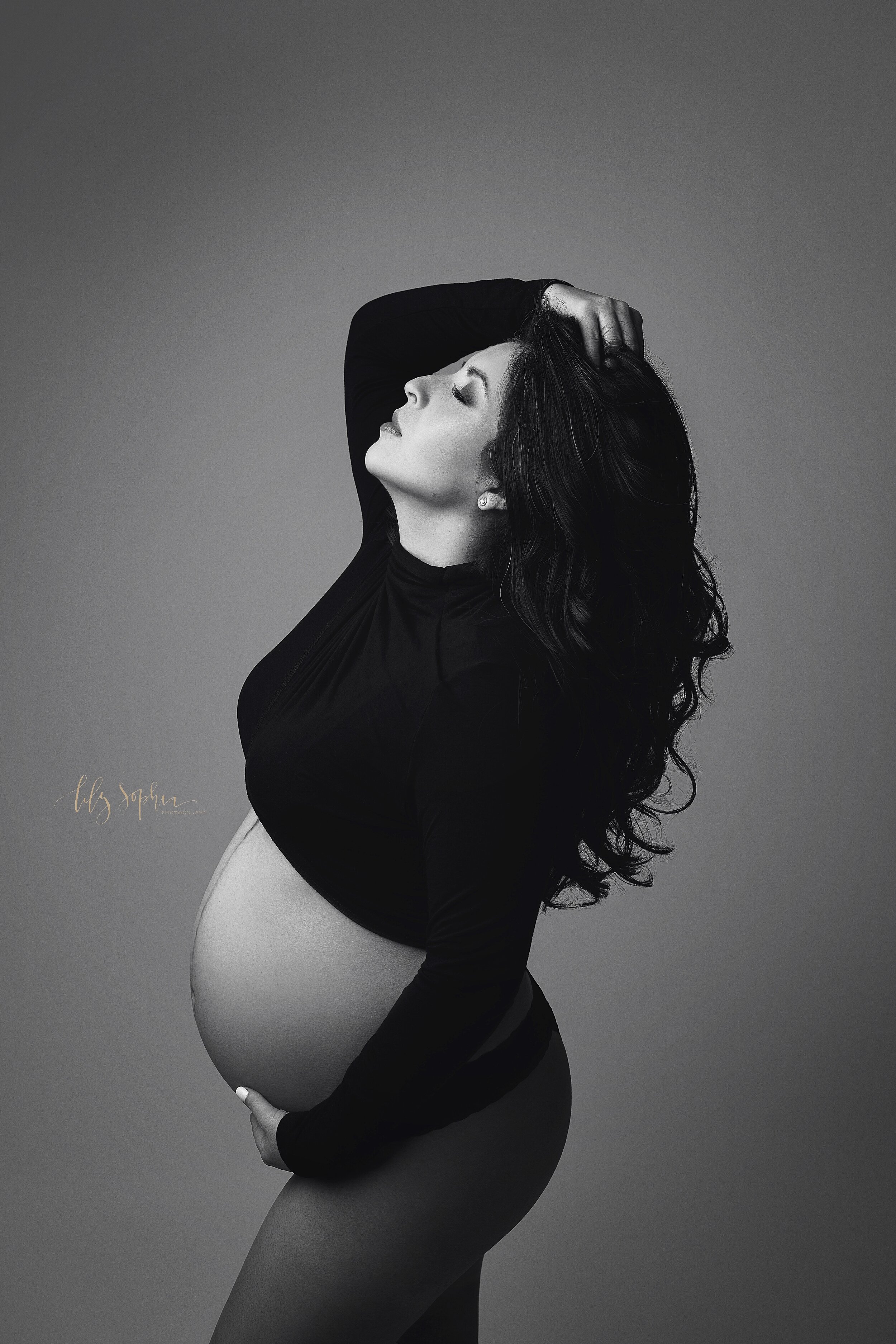  A stunning black and white portrait taken in Atlanta, Georgia of a pregnant Hispanic woman wearing a black long sleeved crop top while running her fingers through her long hair.  