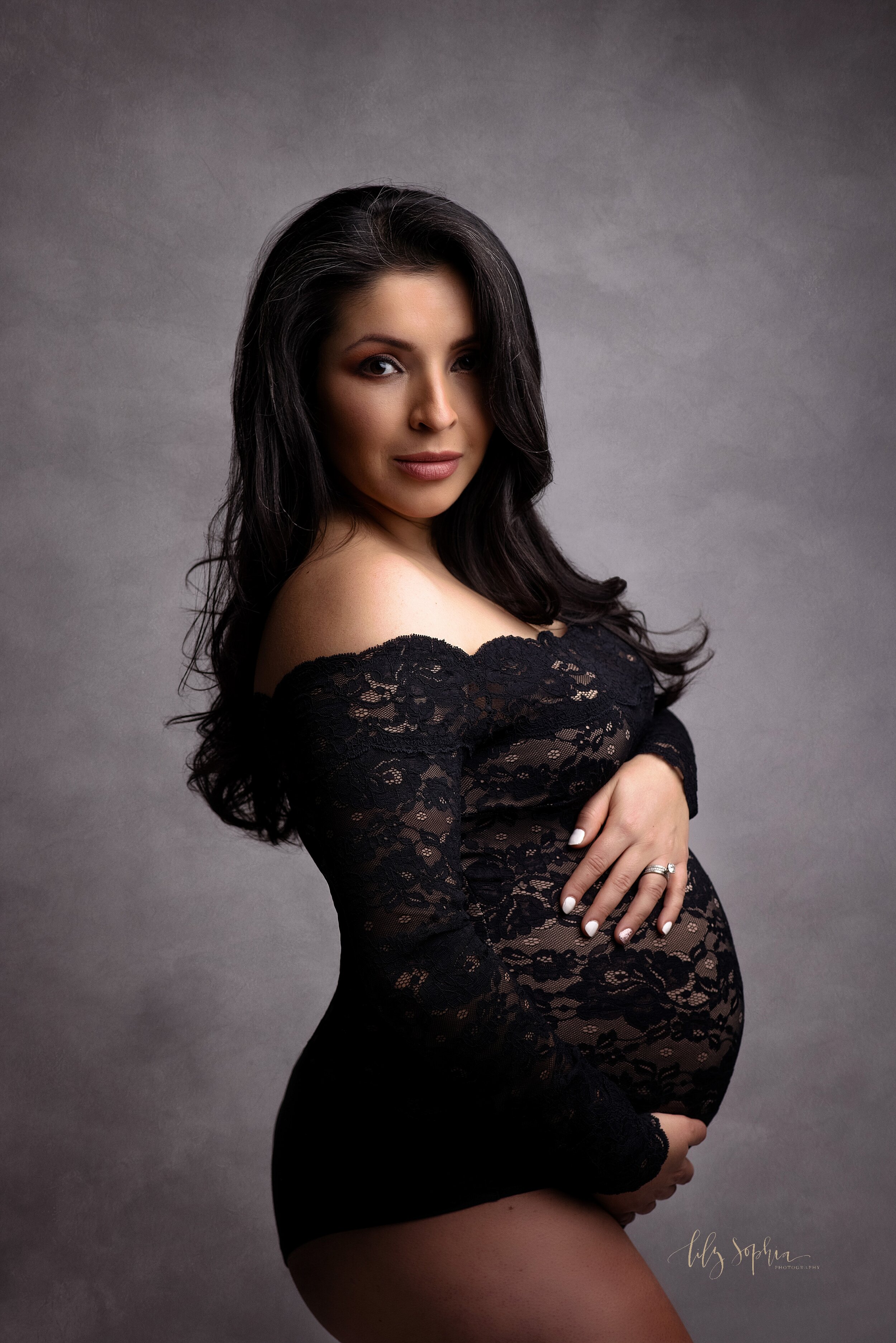  A fine art maternity portrait of a pregnant Hispanic woman with long hair wearing a black off the shoulder lace bodysuit. She is cradling her pregnant belly and gazing serenely at the camera. 