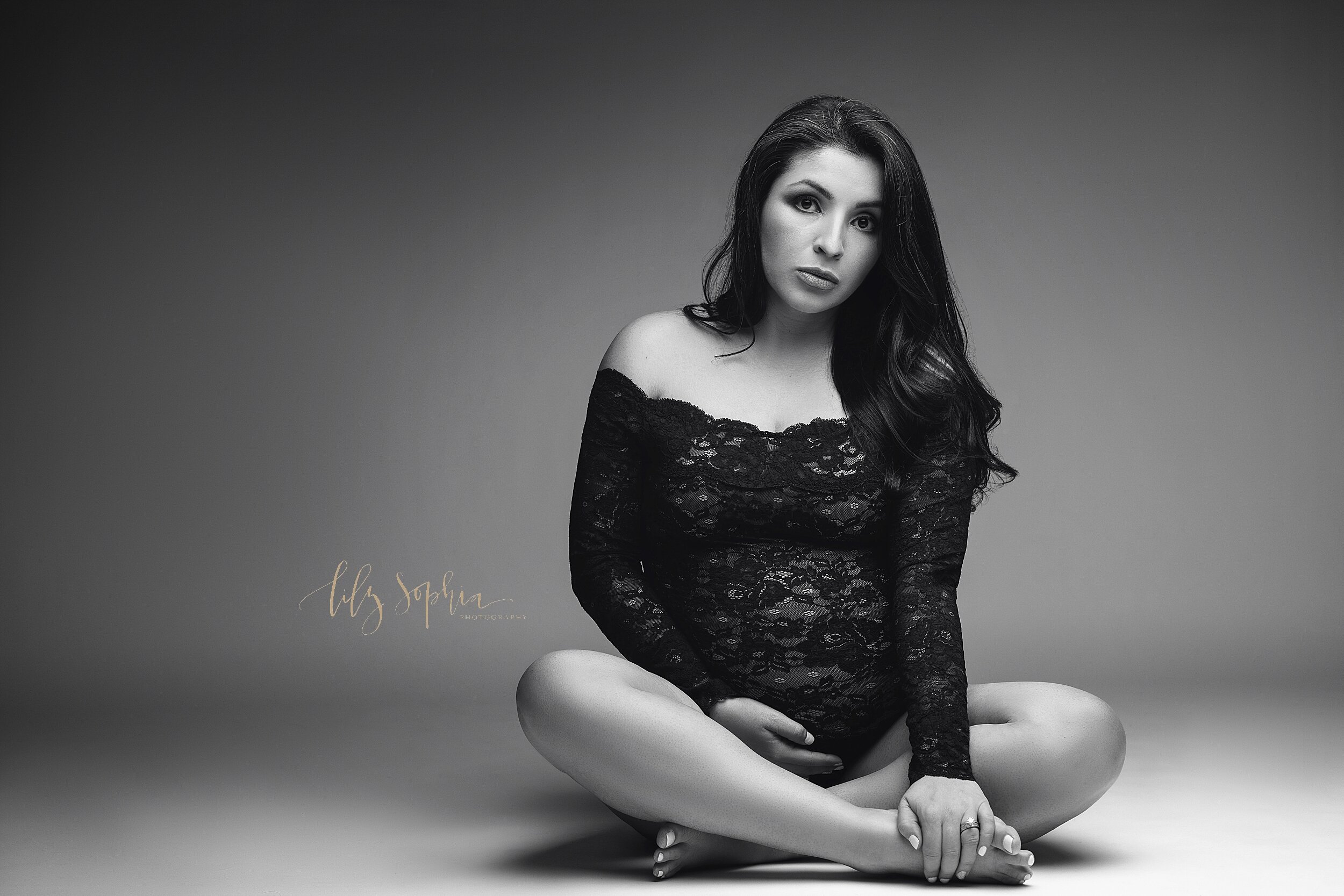  A flack and white fine art maternity portrait of a pregnant Hispanic woman with long hair wearing a black off the shoulder lace bodysuit while sitting on the floor cross legged.  
