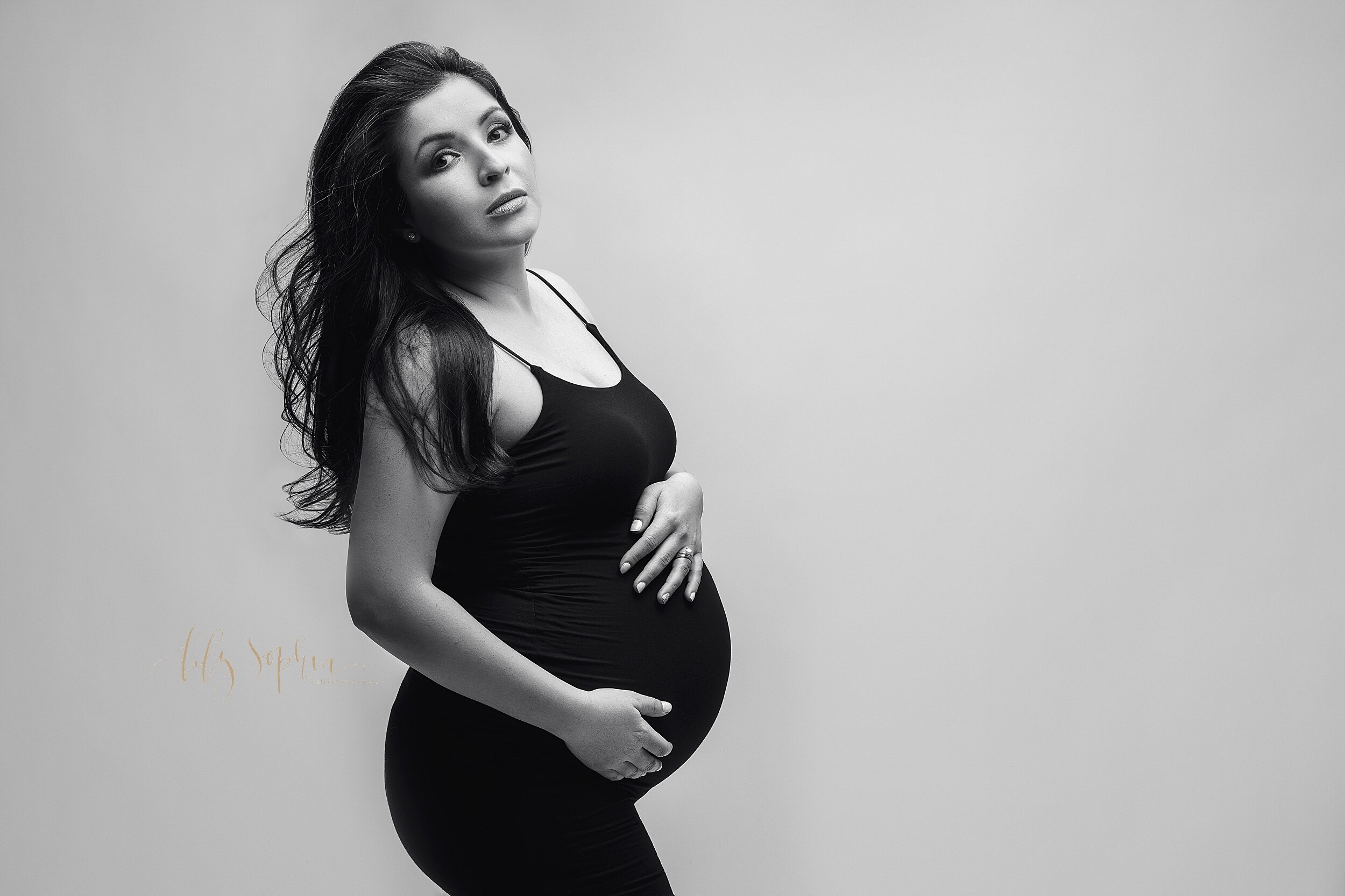  A black and white fine art maternity photo of a pregnant Hispanic woman with long hair blowing in the wind and wearing a black body con dress with spaghetti straps. She is cradling her pregnant belly and has a powerful gaze.  