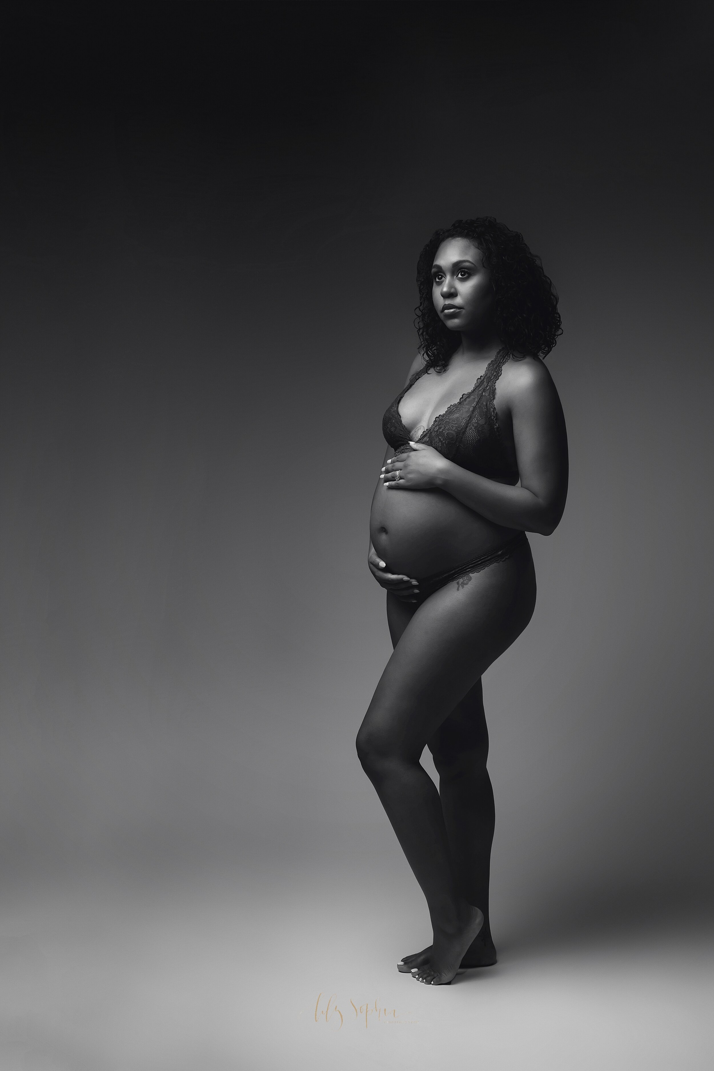  Black and white image of African American expectant woman cradling her pregnant belly wearing lace lingerie. 