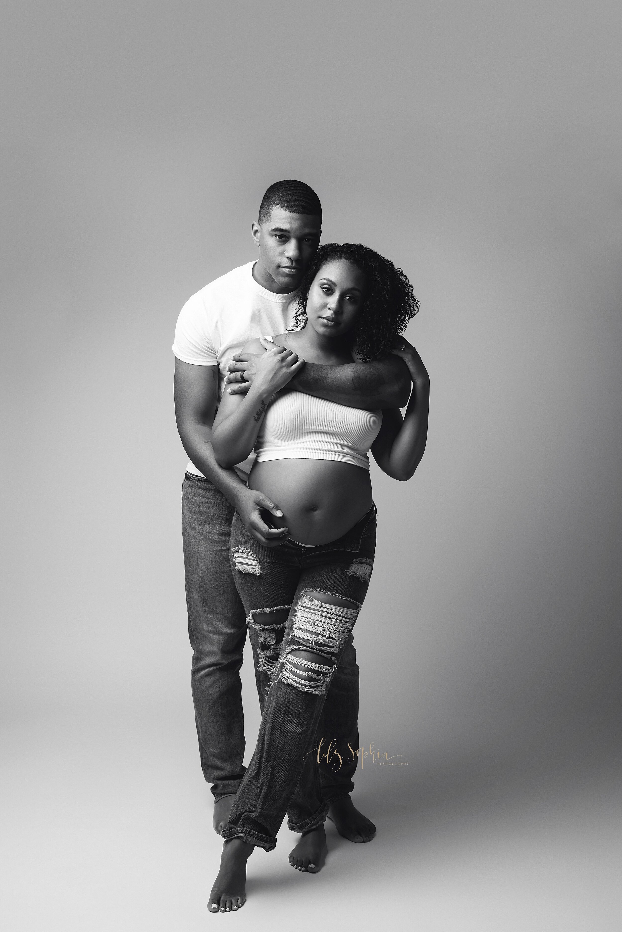  Black and white maternity image of an expectant African American couple embracing while wearing jeans and white tops.  