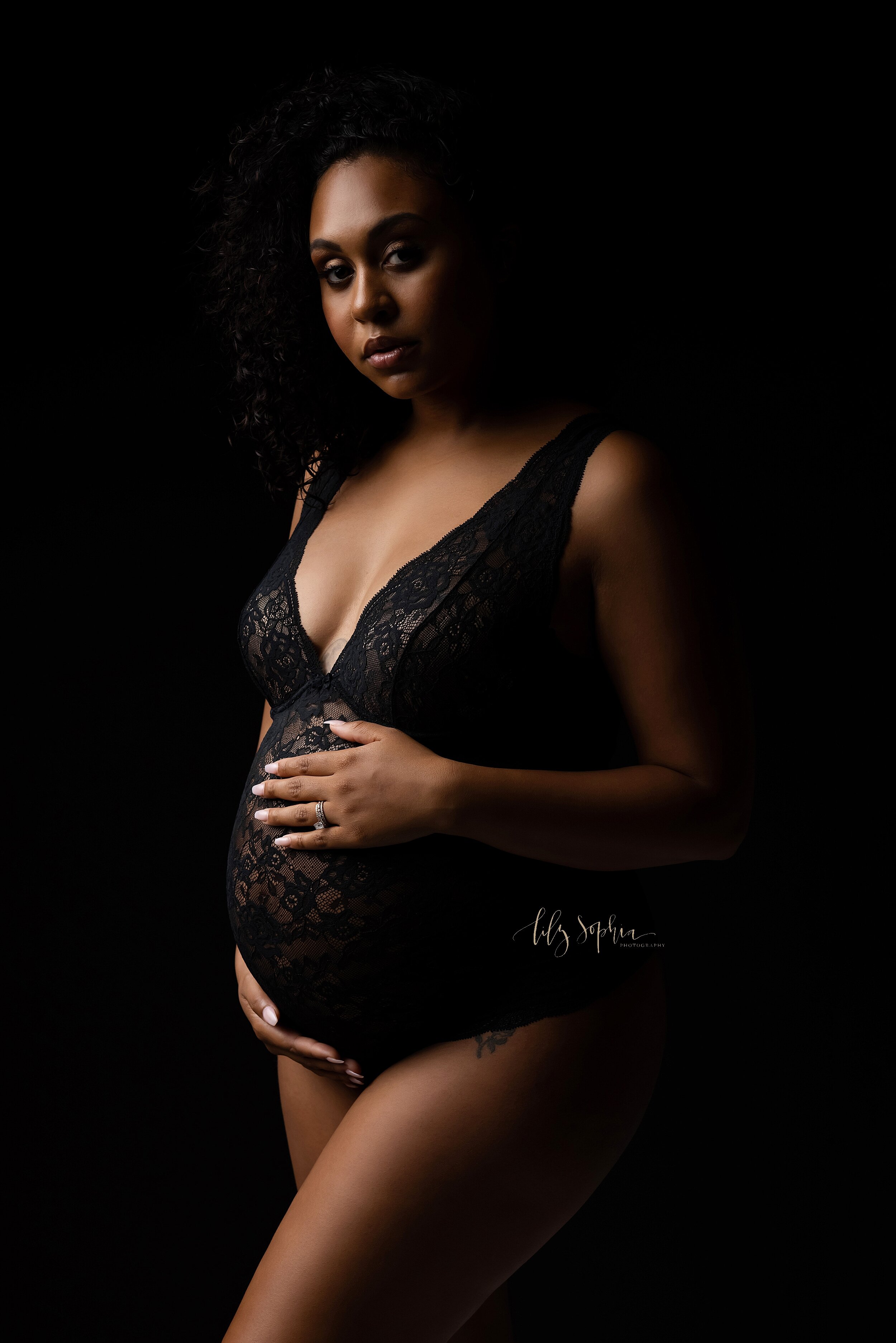  Dramatic fine art modern maternity image of a pregnant black woman with curly hair wearing a lace bodysuit, cradling her baby bump while looking serenely and confidently at the camera. Photo by Lily Sophia Photography located in Atlanta, Georgia.  
