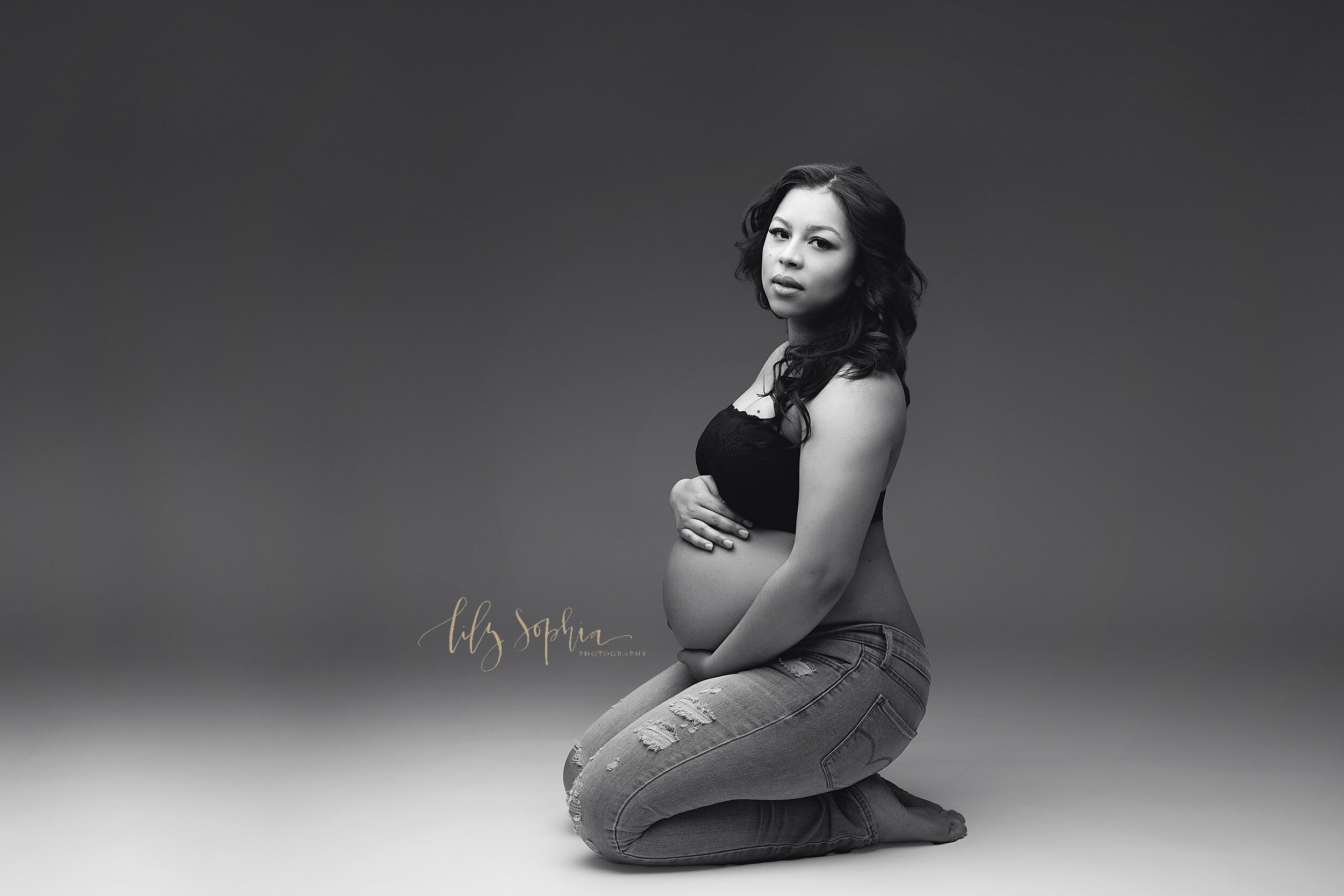  Fine Art Maternity image of Hispanic woman  sitting on her knees wearing a black lace bandeau bra and ripped jeans while cradling her pregnant belly in Atlanta, Georgia studio. 
