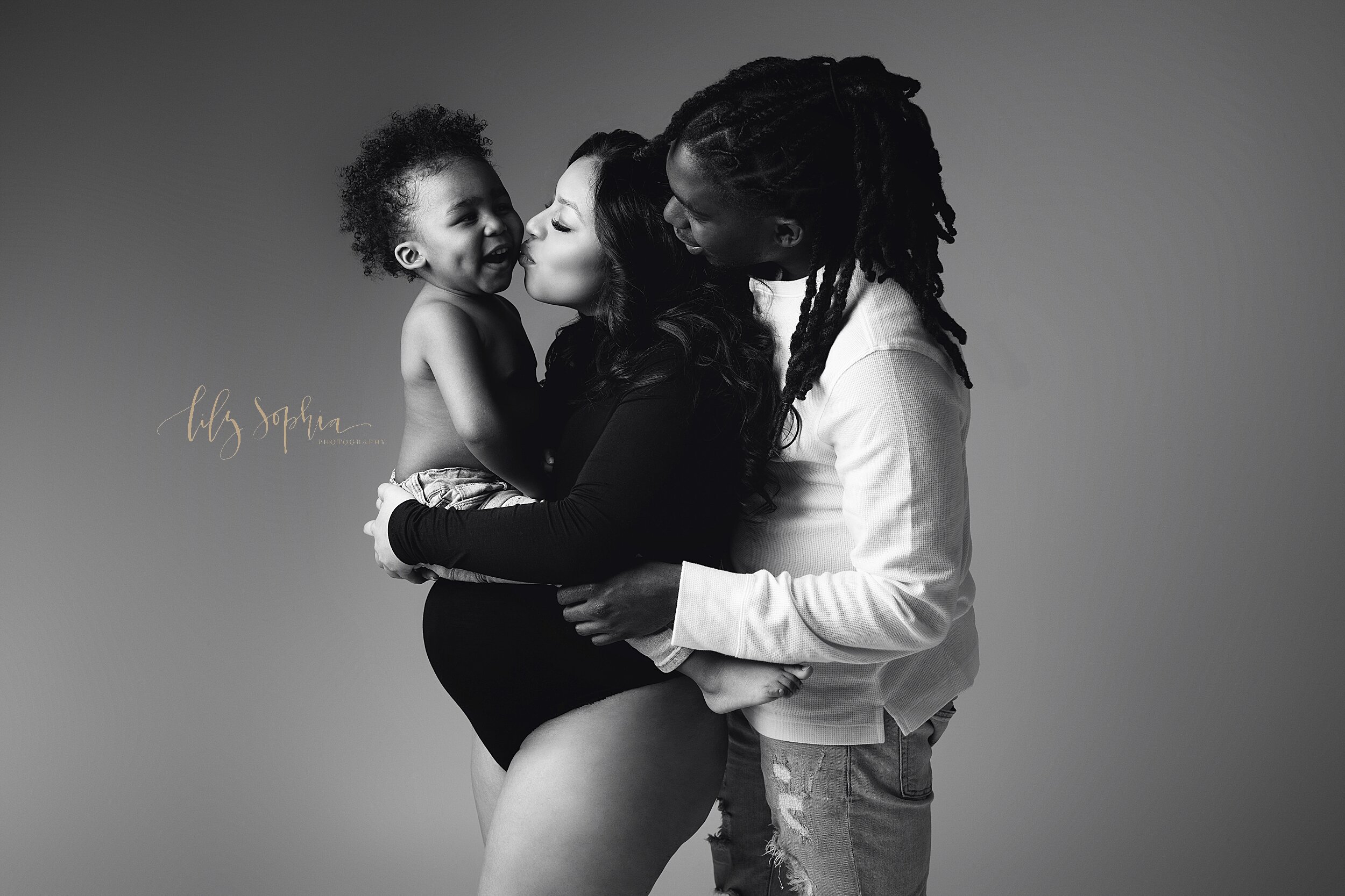  Hispanic mother and black father snuggle with their toddler son in this studio maternity photo. Mother is kissing her son is wearing a black turtleneck bodysuit with her son sitting atop her pregnant belly, with dad in a white sweater looking over m