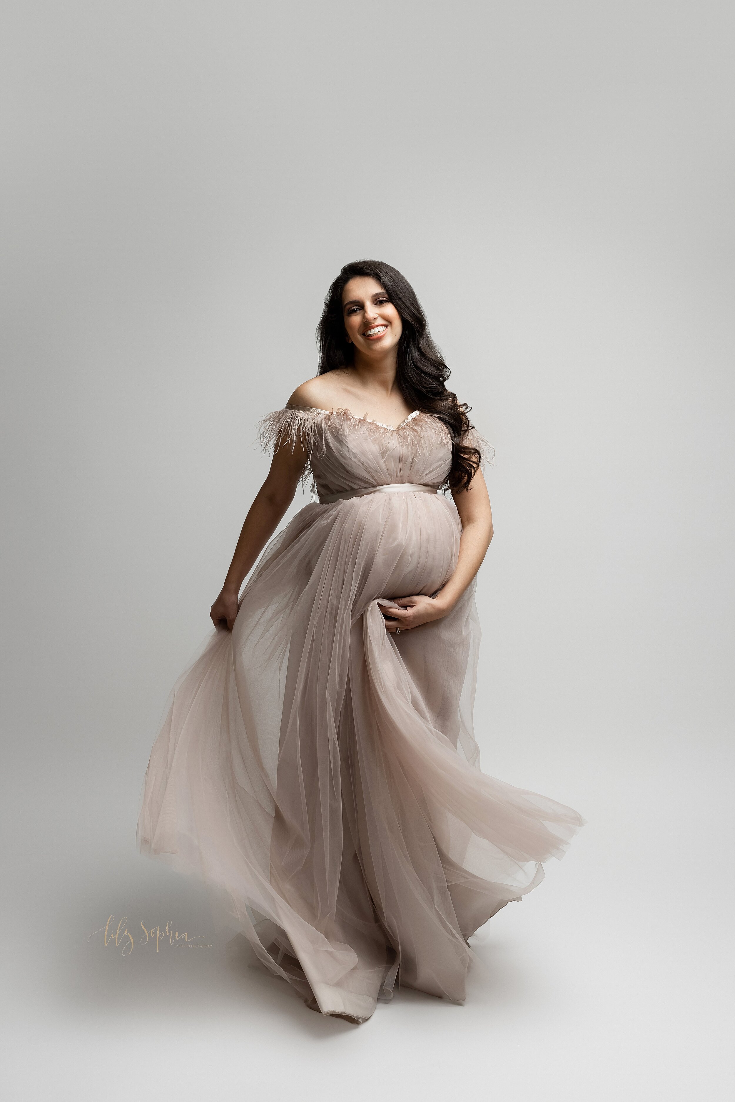  A fine art maternity photo of a happy and smiling pregnant woman with long brown hair wearing a grey tulle haute couture maternity gown with ostrich feathers with one hand on her belly and the other holding the dress. 