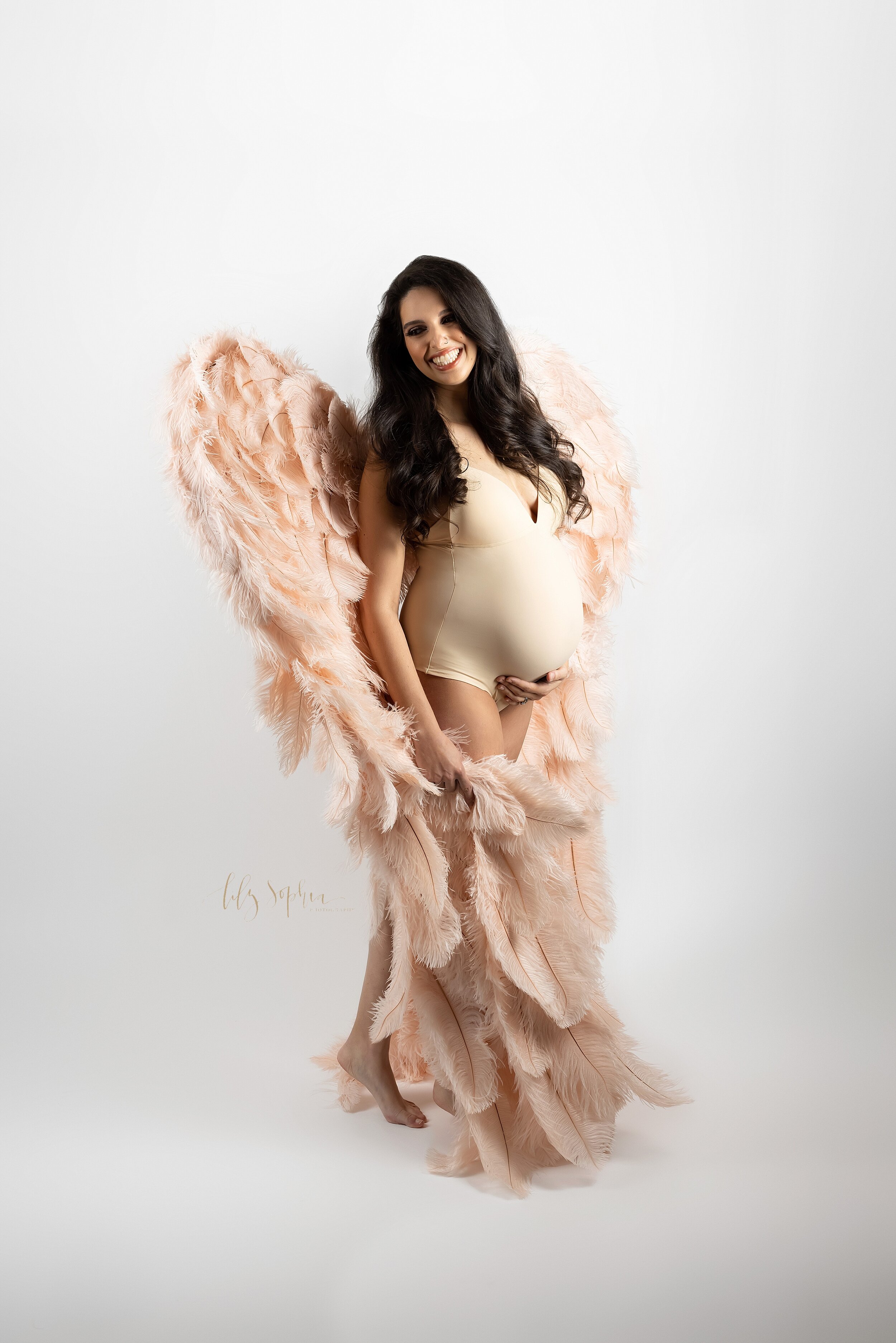  Atlanta fine art modern maternity photo of happy pregnant woman with a big smile and long brown hair wearing a nude bodysuit and blush pink feather angel wings inspired by the Victoria’s Secret runway.  