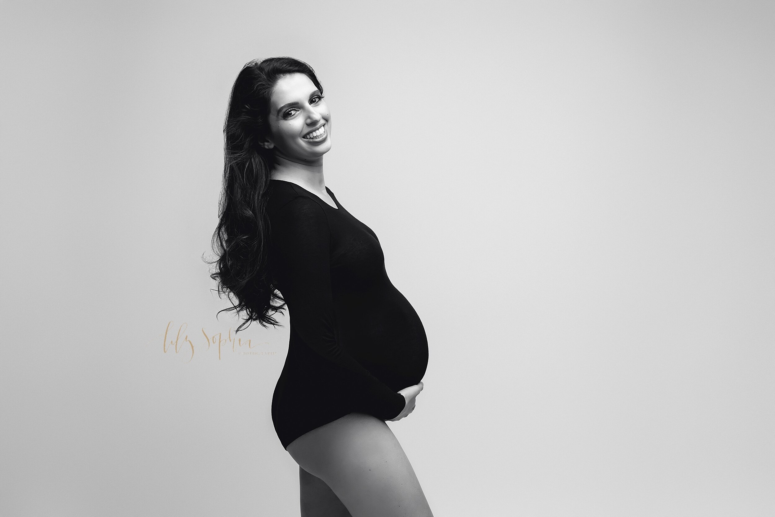  A b&amp;w fine art modern maternity photo of a happy pregnant woman with long brown hair wearing a simple black bodysuit and looking at the camera in the Atlanta, GA studio of Lily Sophia Photography. 
