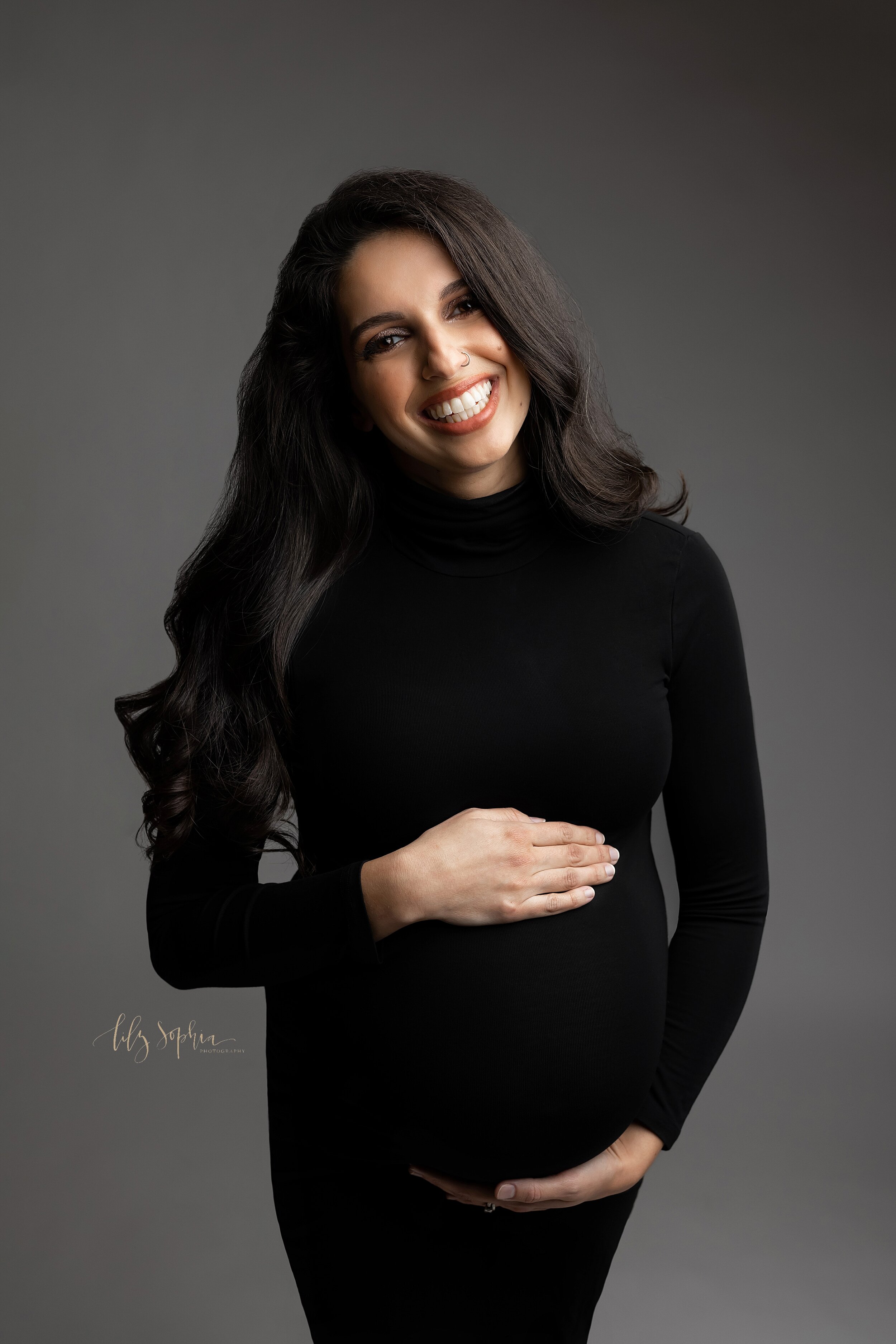  Fine Art modern maternity studio picture of happy smiling pregnant woman with long curly brown hair wearing black mock neck body con dress and cradling her baby bump in Atlanta, Georgia.  