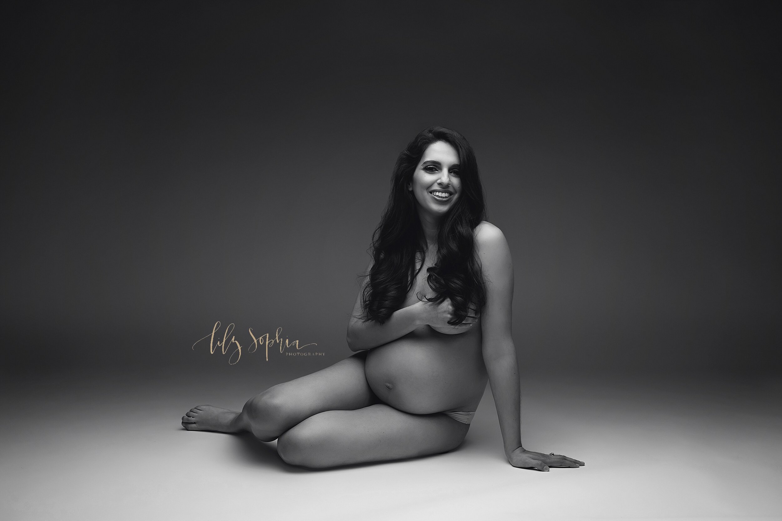  A B&amp;W fine art modern maternity studio portrait of a happy pregnant woman with long brown hair sitting like a mermaid on the floor while covering her breasts. 