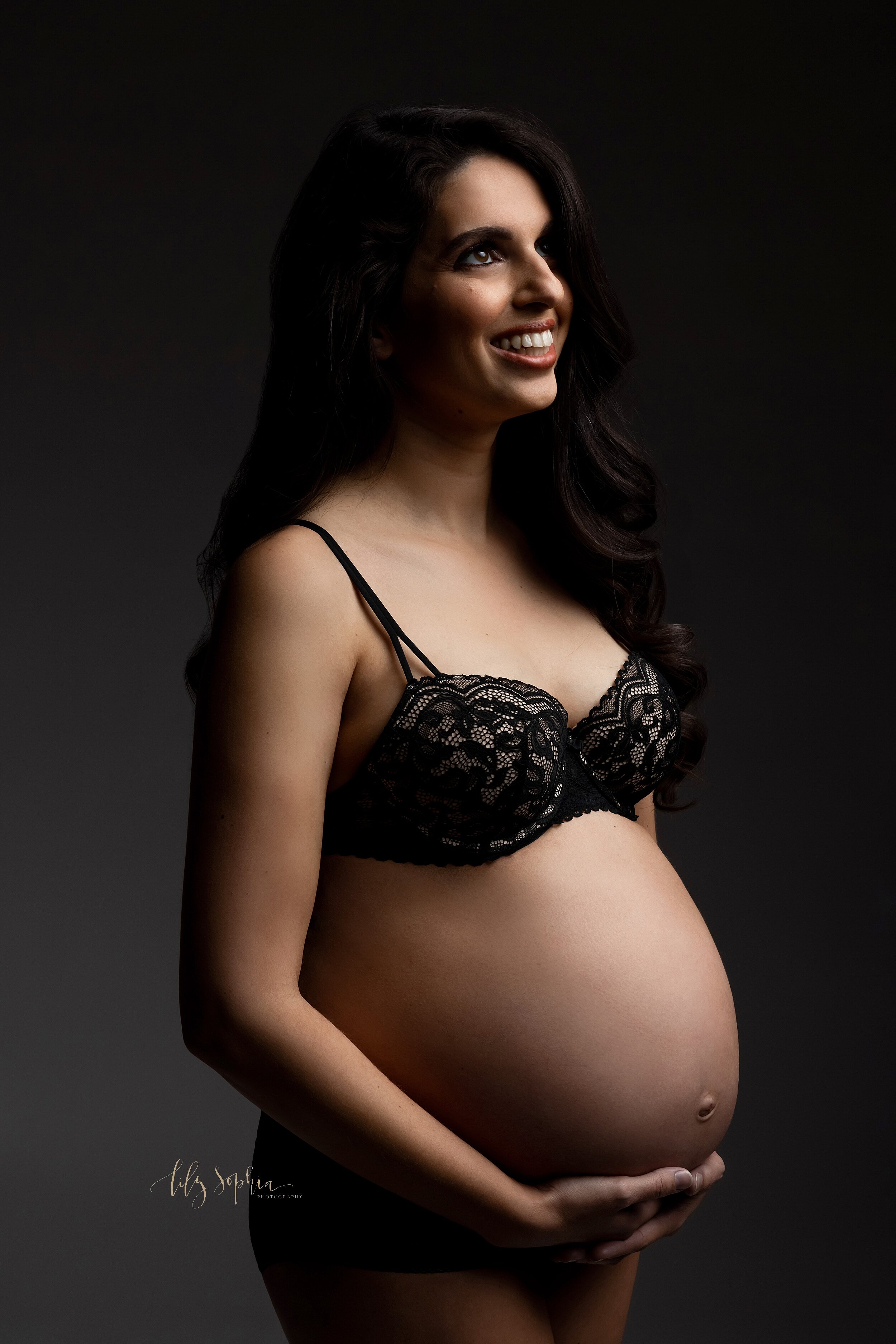  A fine art modern maternity pic of a happy pregnant woman cradling her baby bump while wearing a black lace bra in Atlanta. 