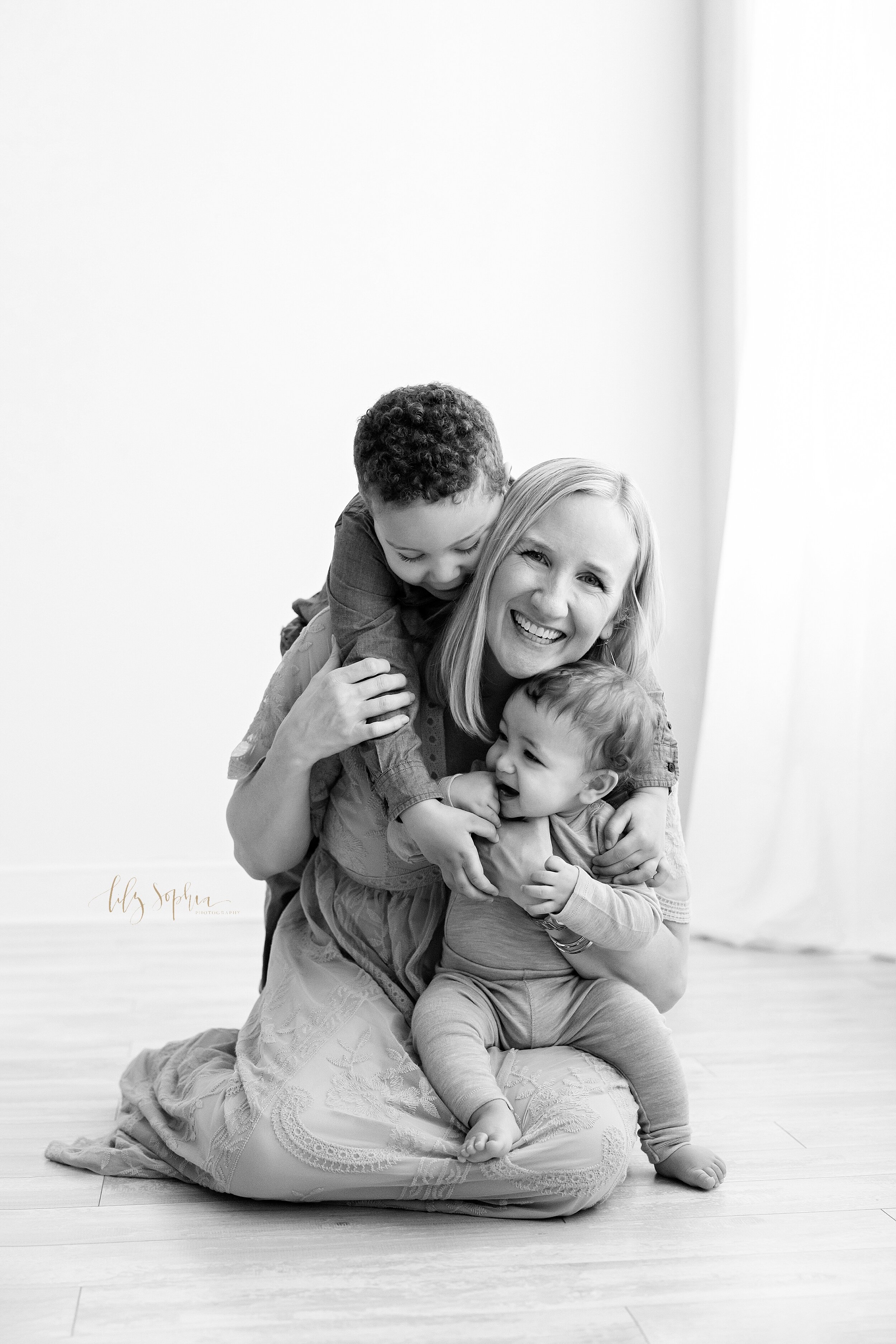  Black and white family image of a mom sitting on the floor of a studio with her toddler son climbing on her back and her seven month old son sitting on her lap taken near Morningside in Atlanta. 