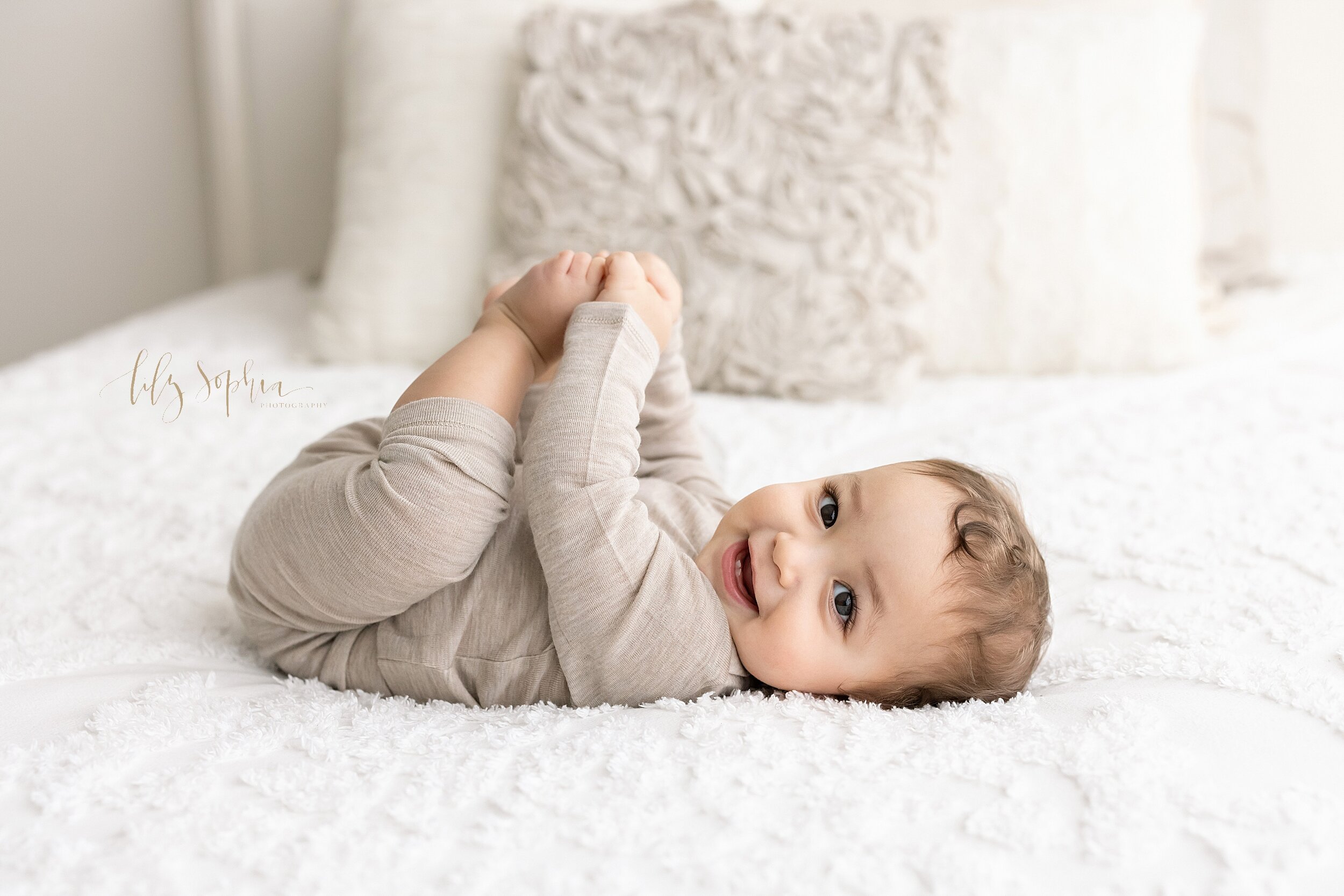  Milestone photo of a smiling seven month old baby boy as he lies on a bed on his back and plays with his toes taken in a studio in the Ponce City Market area of Atlanta in natural light. 