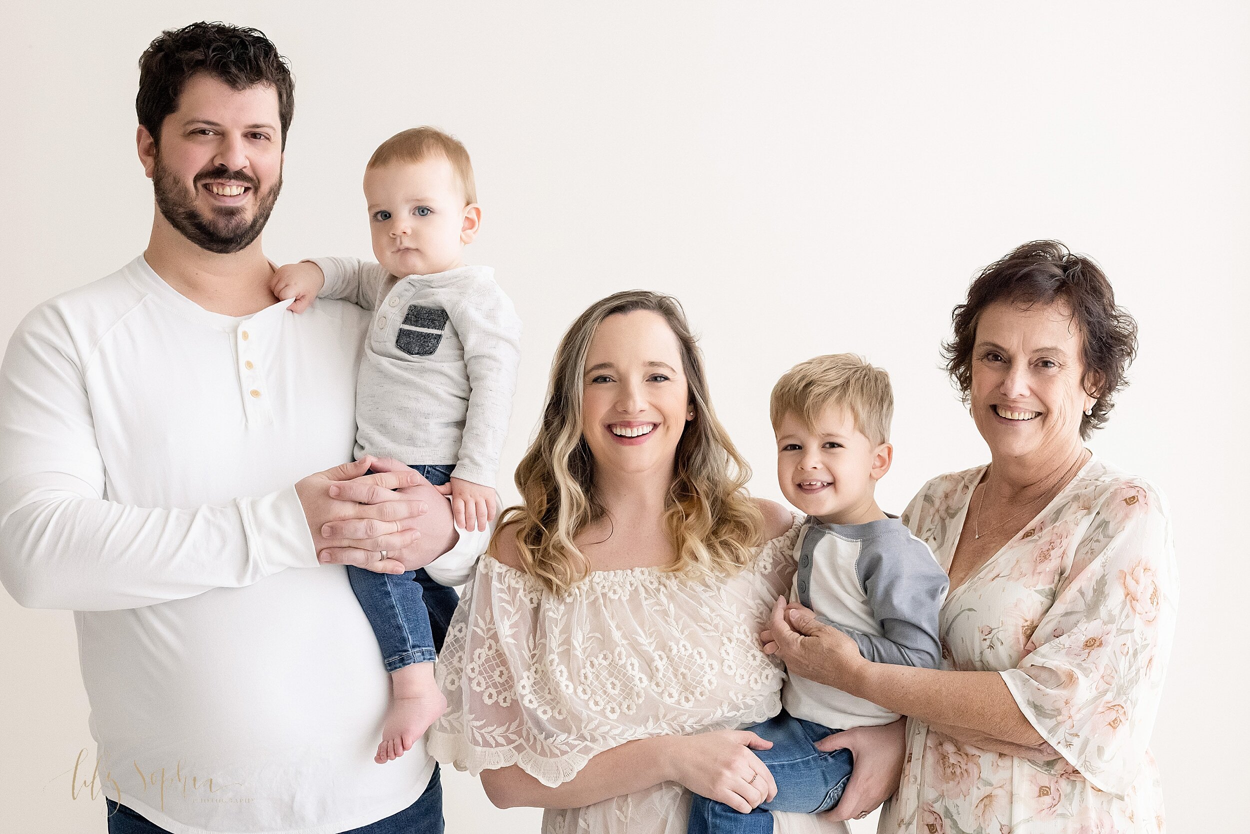  Intergenerational family portrait of a father holding his one-year old son, his wife holding their toddler son, and grandma standing next to them and wrapping her arms around her toddler grandson as they stand in a natural light studio in the Poncey