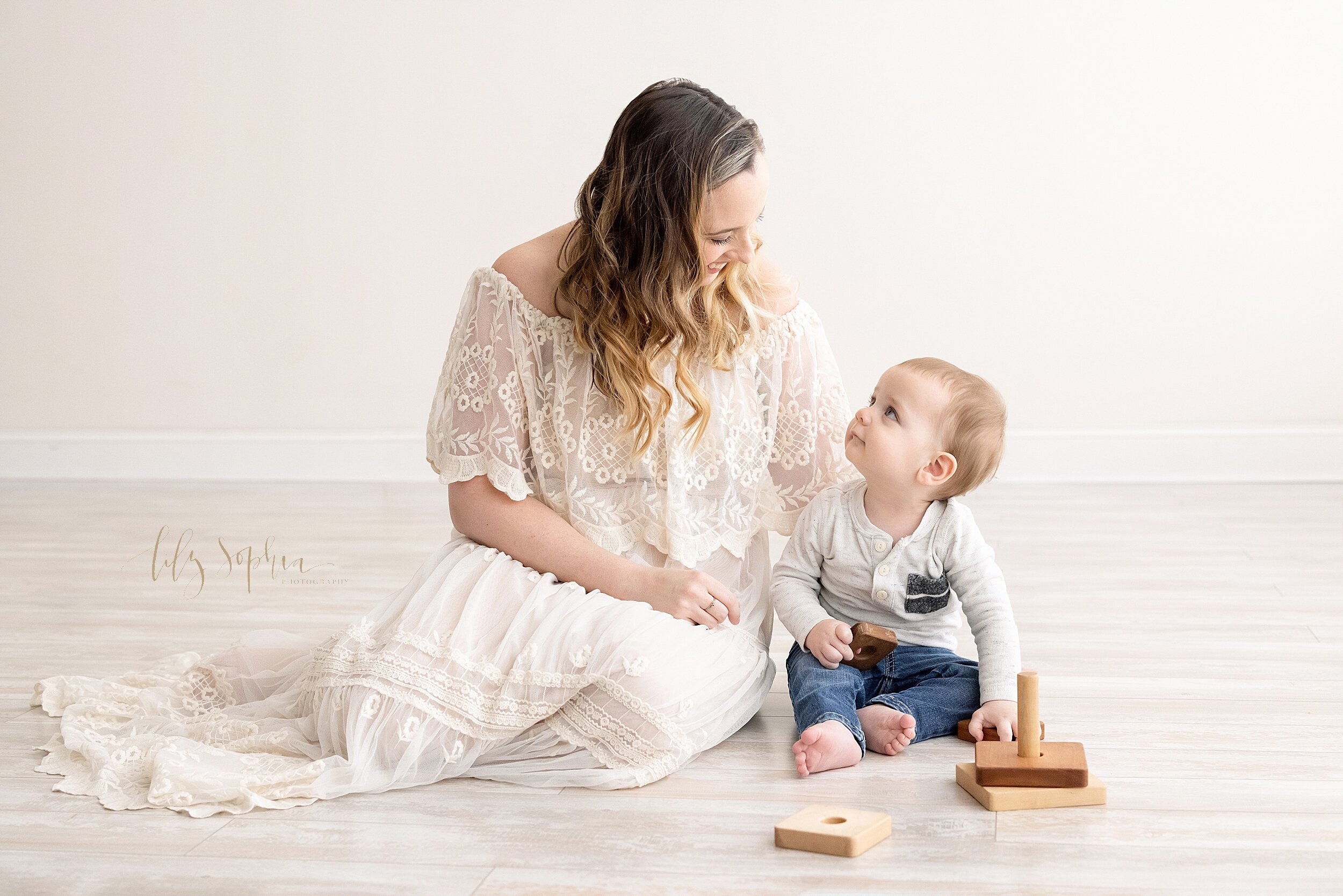  Family photo of a one-year old boy as he sits next to his mom on the floor of a natural light studio in the Buckhead area of Atlanta, Georgia and plays with a wooden stacking toy while looking of into his mother’s eyes. 