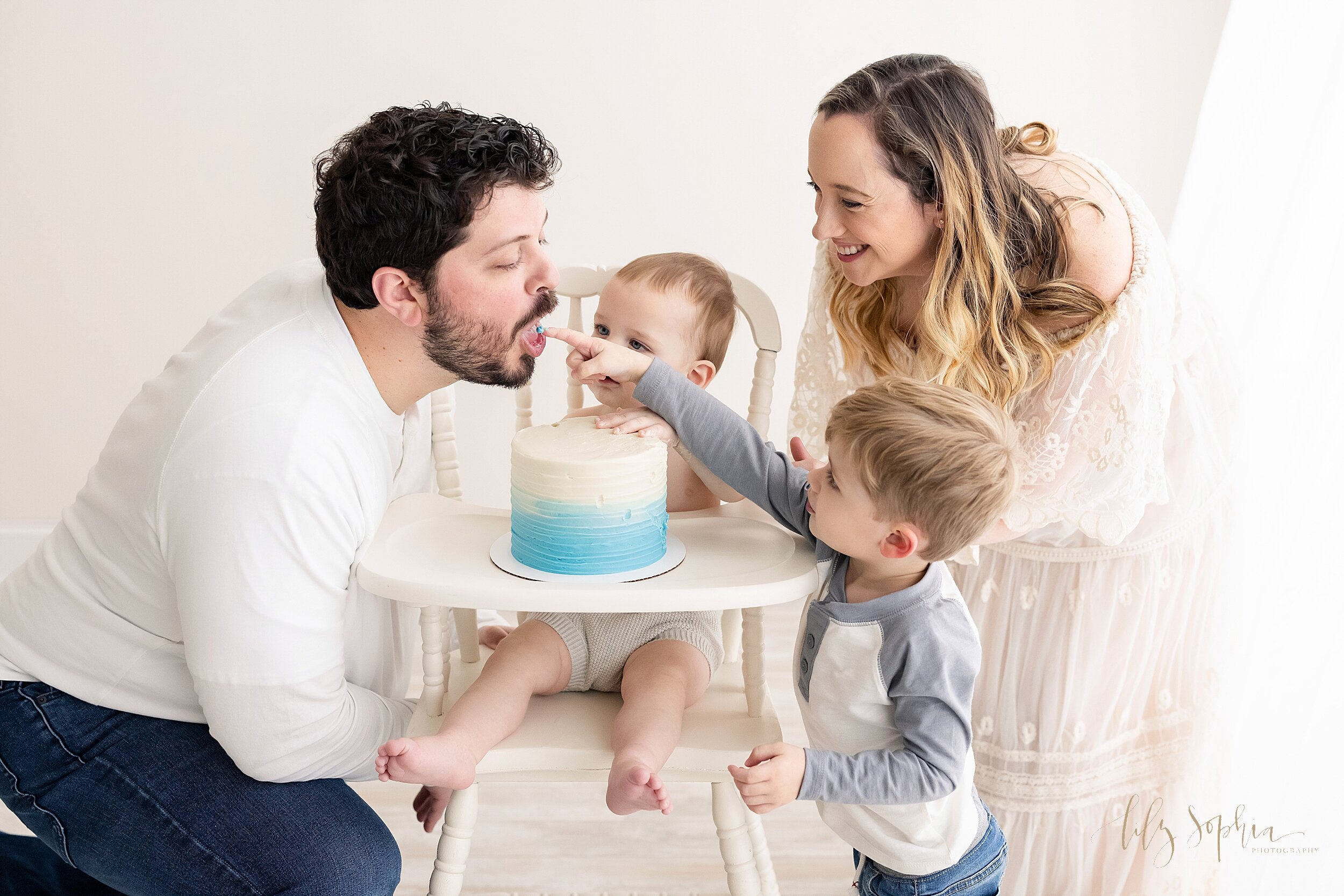  Family photo of the first birthday of little boy who is sitting with his cake on the tray of an antique high chair with his father on his left side and his mother and toddler brother on his right side as his toddler brother sticks his finger with ic