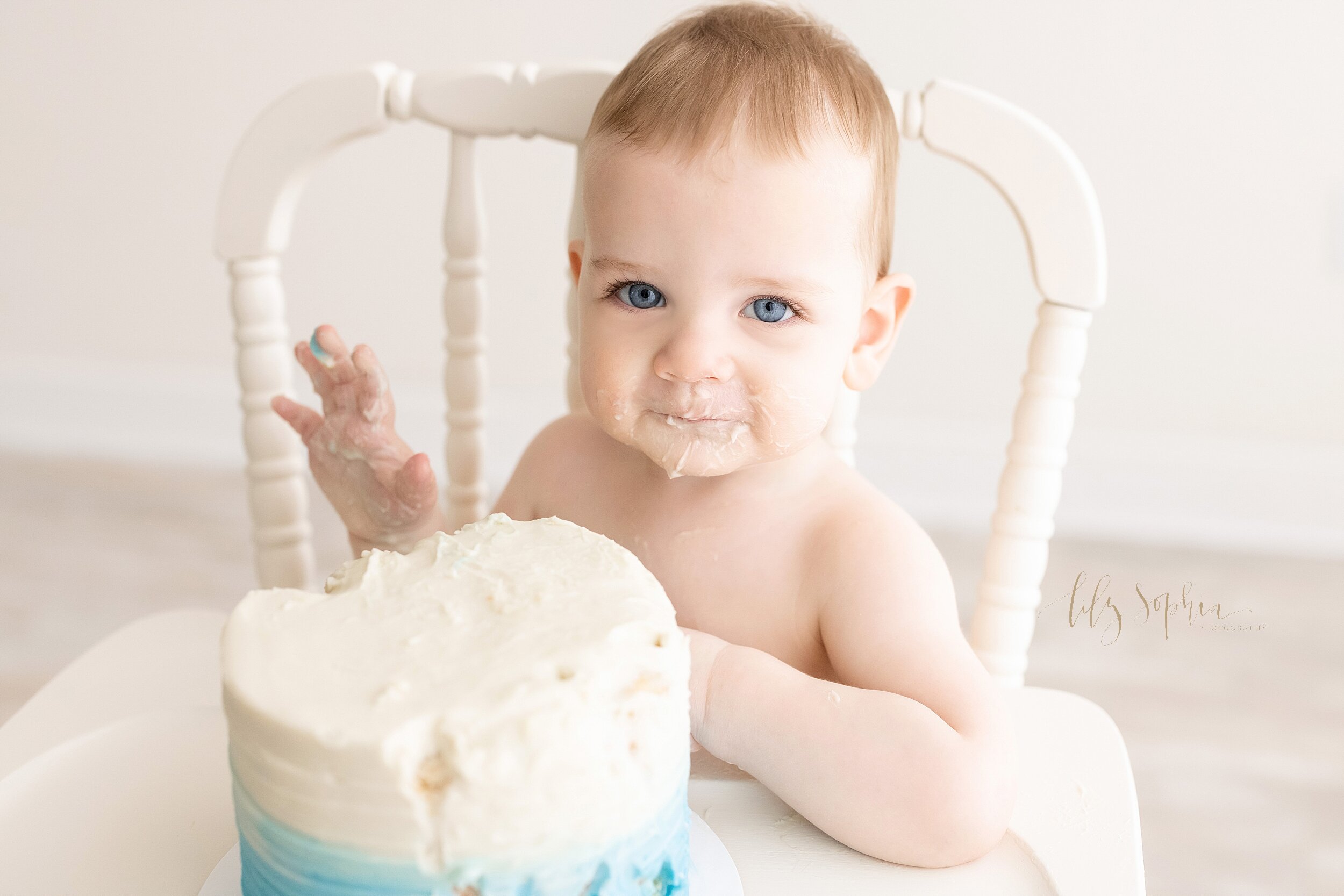  First birthday photo of a one-year old baby boy with his hands and face covered with icing from his smash cake as he sits smiling in an antique highchair in a natural light studio in the Vinings area of Atlanta. 