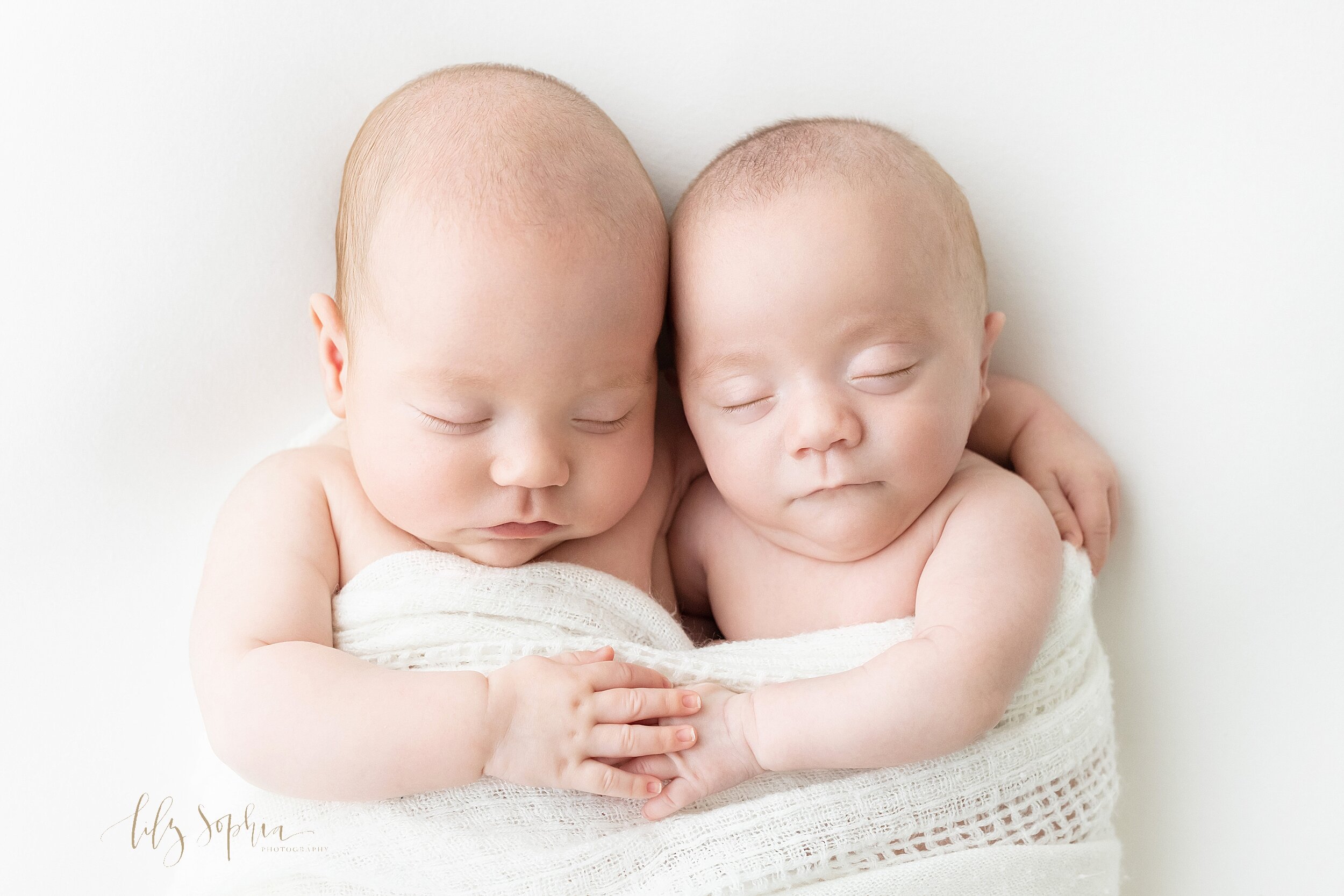  Newborn photograph of twin boys with their arms around each other and their other hands on top of one another as they are swaddled together in one white knitted blanket as they sleep in a studio using natural light near Oakhurst in Atlanta, Georgia.