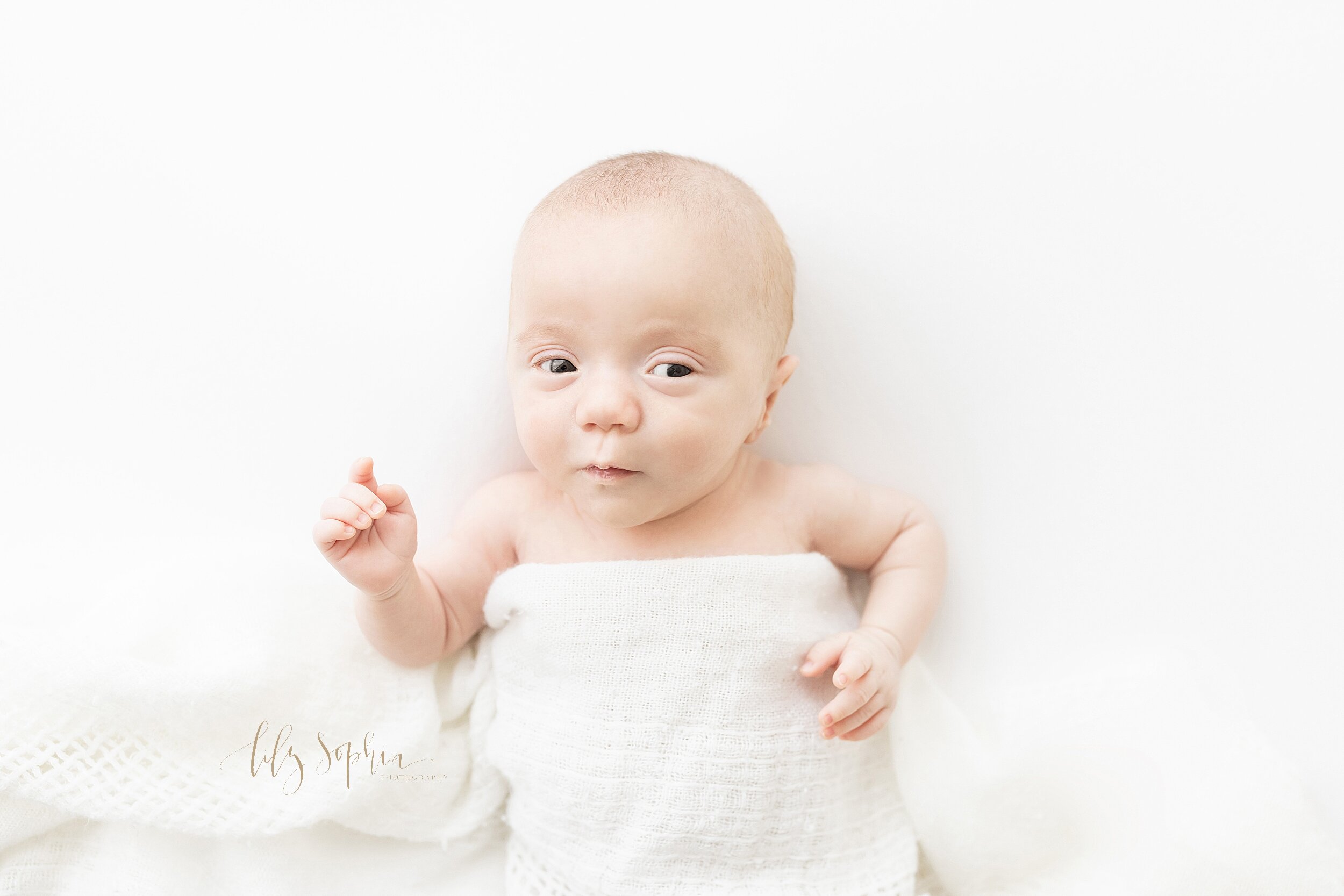  Newborn photograph of an awake infant boy draped with a white knitted blanket as he raises one arm and appears to want to speak taken in a natural light studio near Midtown in Atlanta, Georgia. 
