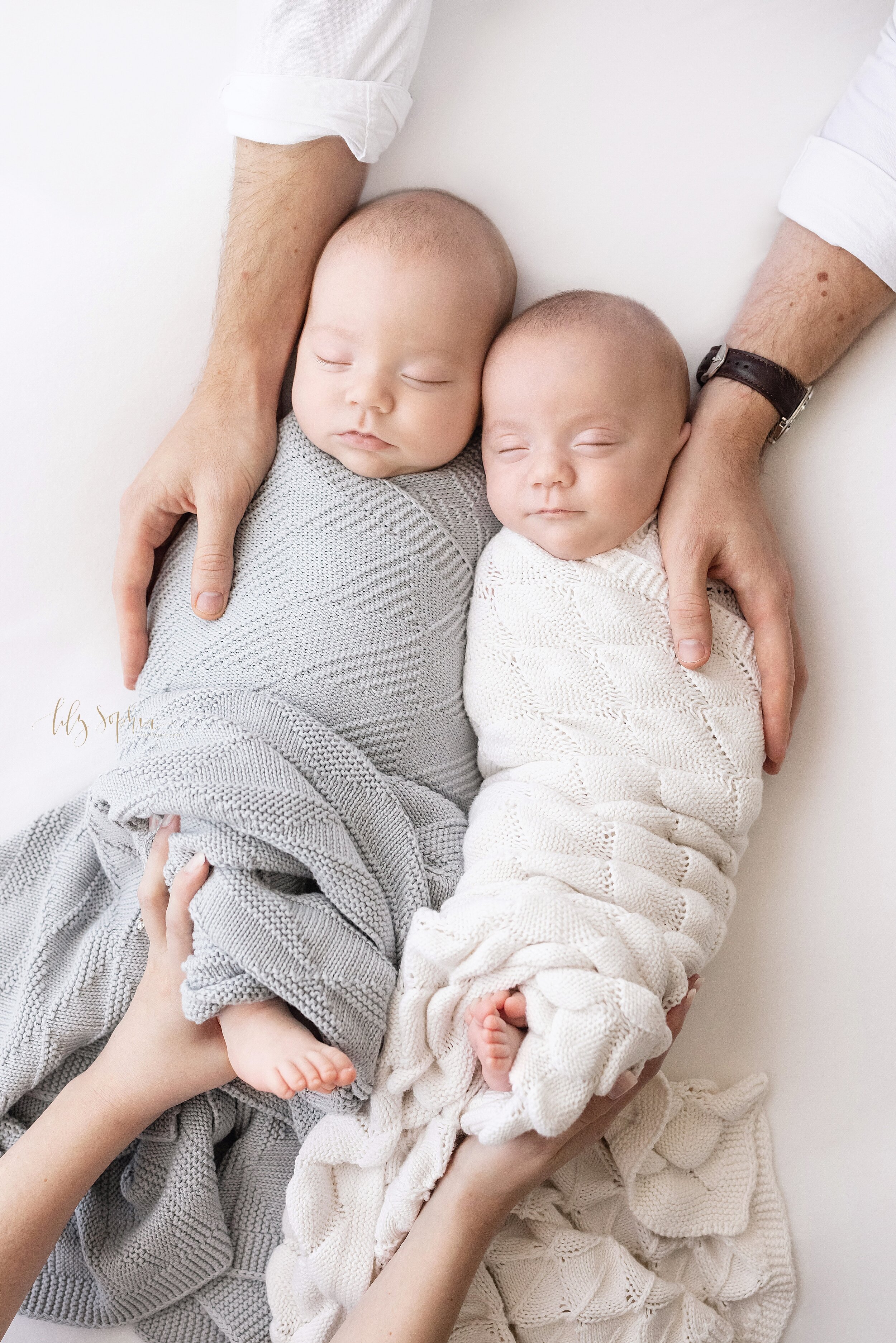  Newborn photo session of twin boys peacefully sleeping next to one another swaddled in knit blankets to their chins with dad placing his hands on the shoulders of the infants and mom placing her hands on their sons’ feet taken in a studio in Ponce C