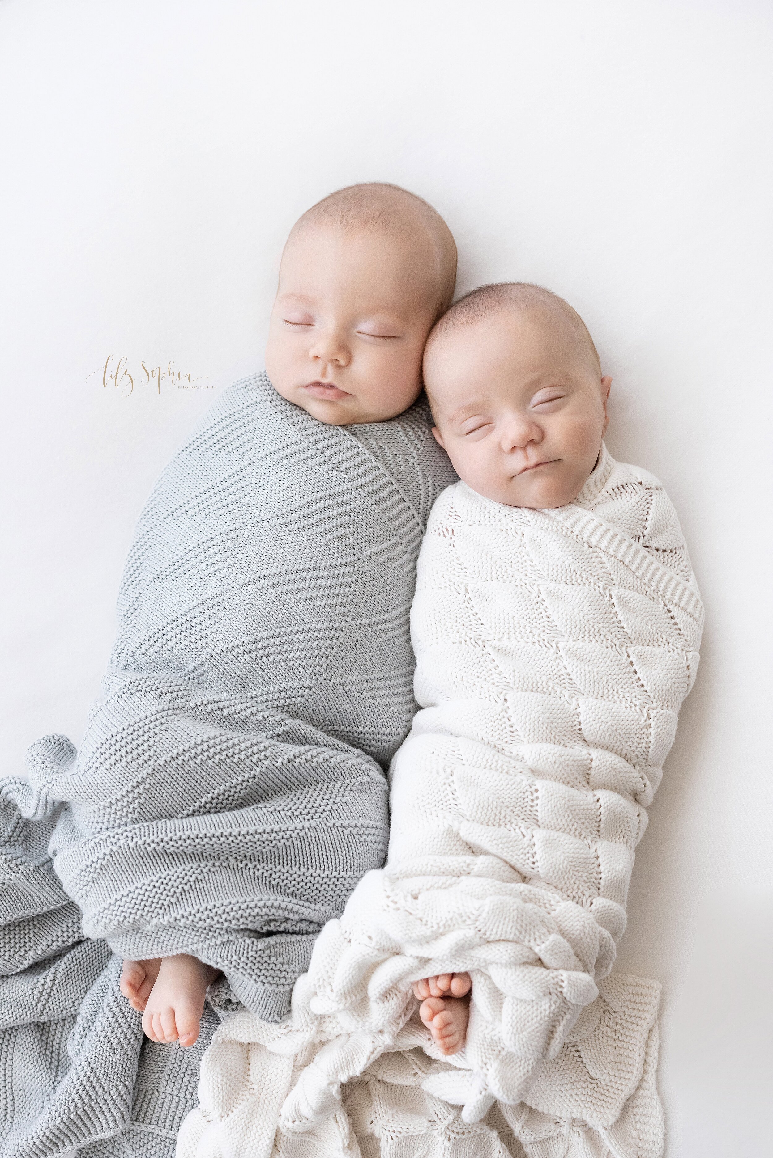 Newborn portrait of peacefully sleeping twin boys swaddled in blankets to their chins with their tiny toes peeking out lying next to one another with one of the twin’s heads touching the cheek of his brother taken in a natural light studio near Ansl