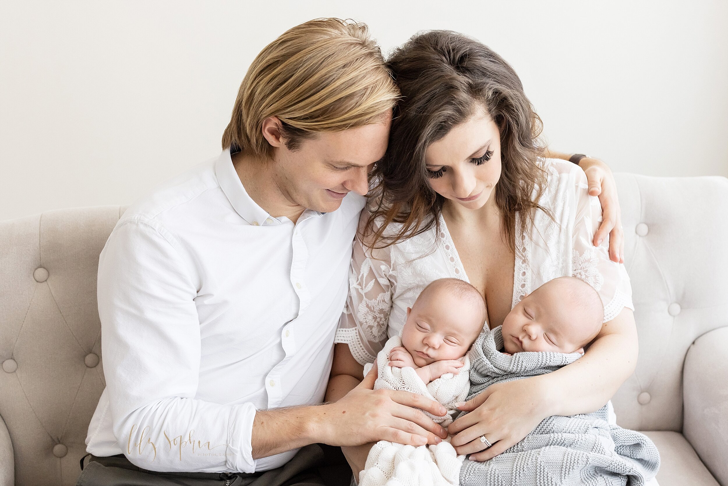  Family photograph of a mother and a father sitting next to one another on a sofa in a natural light studio while dad wraps his left hand around his wife’s shoulder and mom holds their newborn twin boys on her lap and the two of them admire the infan
