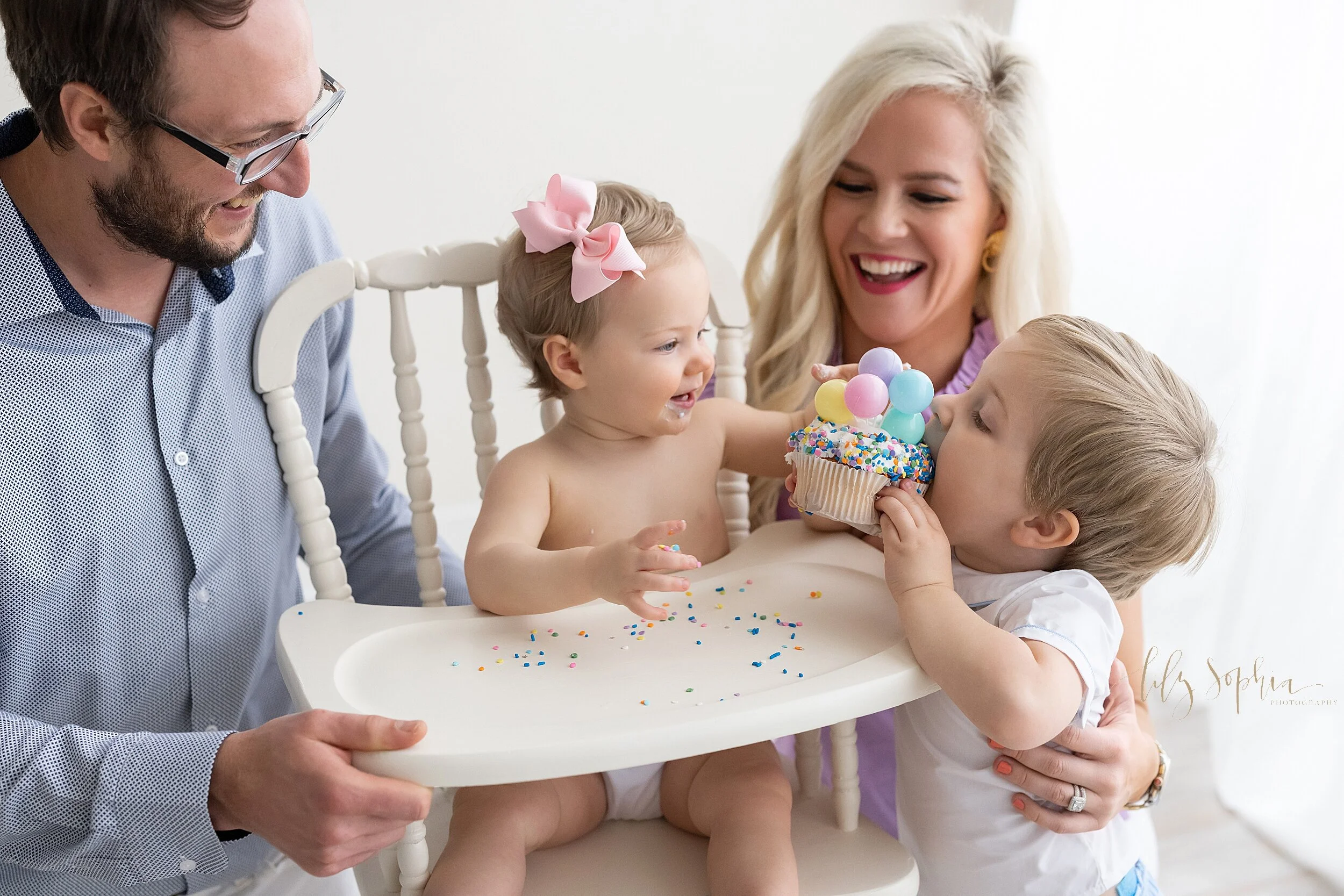  First birthday photo session of little girl wearing a pink bow in her hair as she sits in an antique high chair with her dad on her left and her mom on her right as she feeds her birthday cupcake to her brother taken in a natural light studio near A