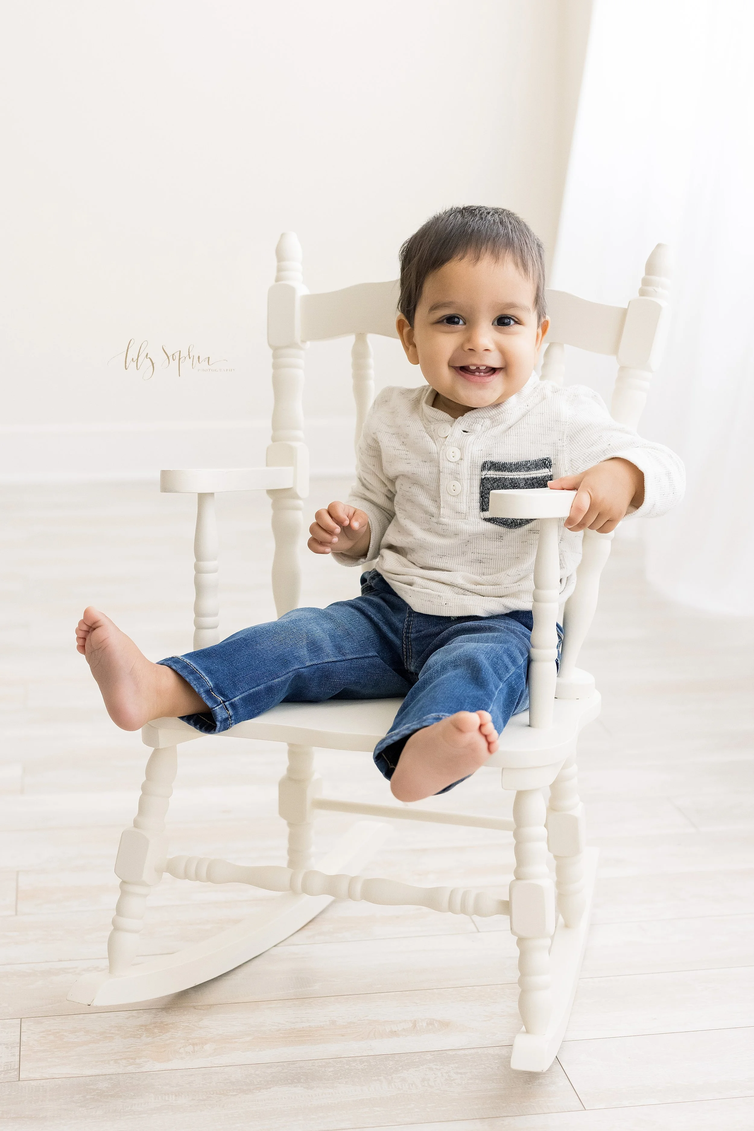  First birthday picture of a smiling one year old dark haired and dark eyed boy as he sits in a white rocking chair in front of a window streaming natural light in a studio near Poncey Highlands in Atlanta, Georgia. 
