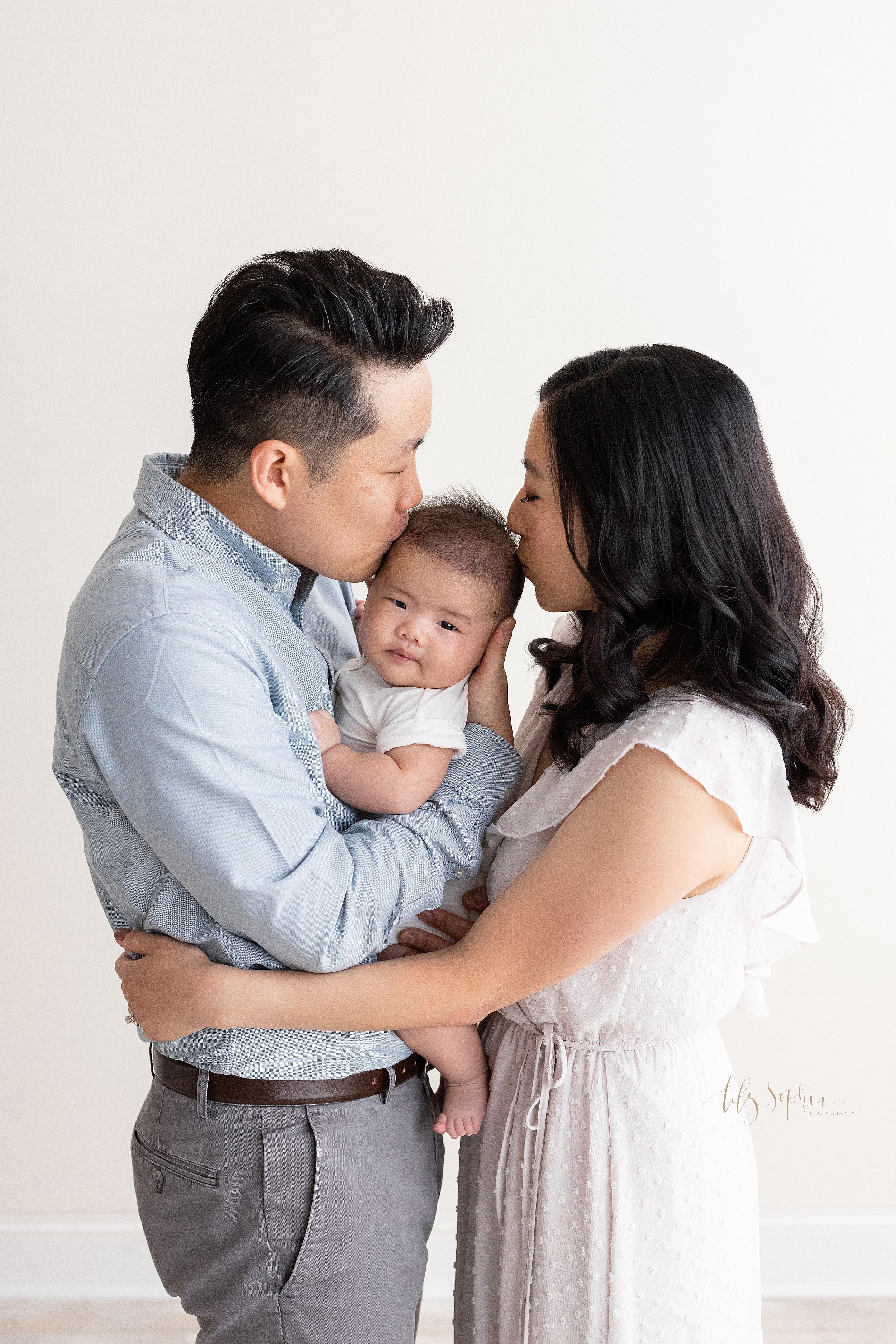  Family portrait of an Asian family as they celebrate one hundred days with their son with dad holding his son against his chest and kissing the right side of his son’s head and mom facing dad and kissing the left side of their son’s head taken in a 