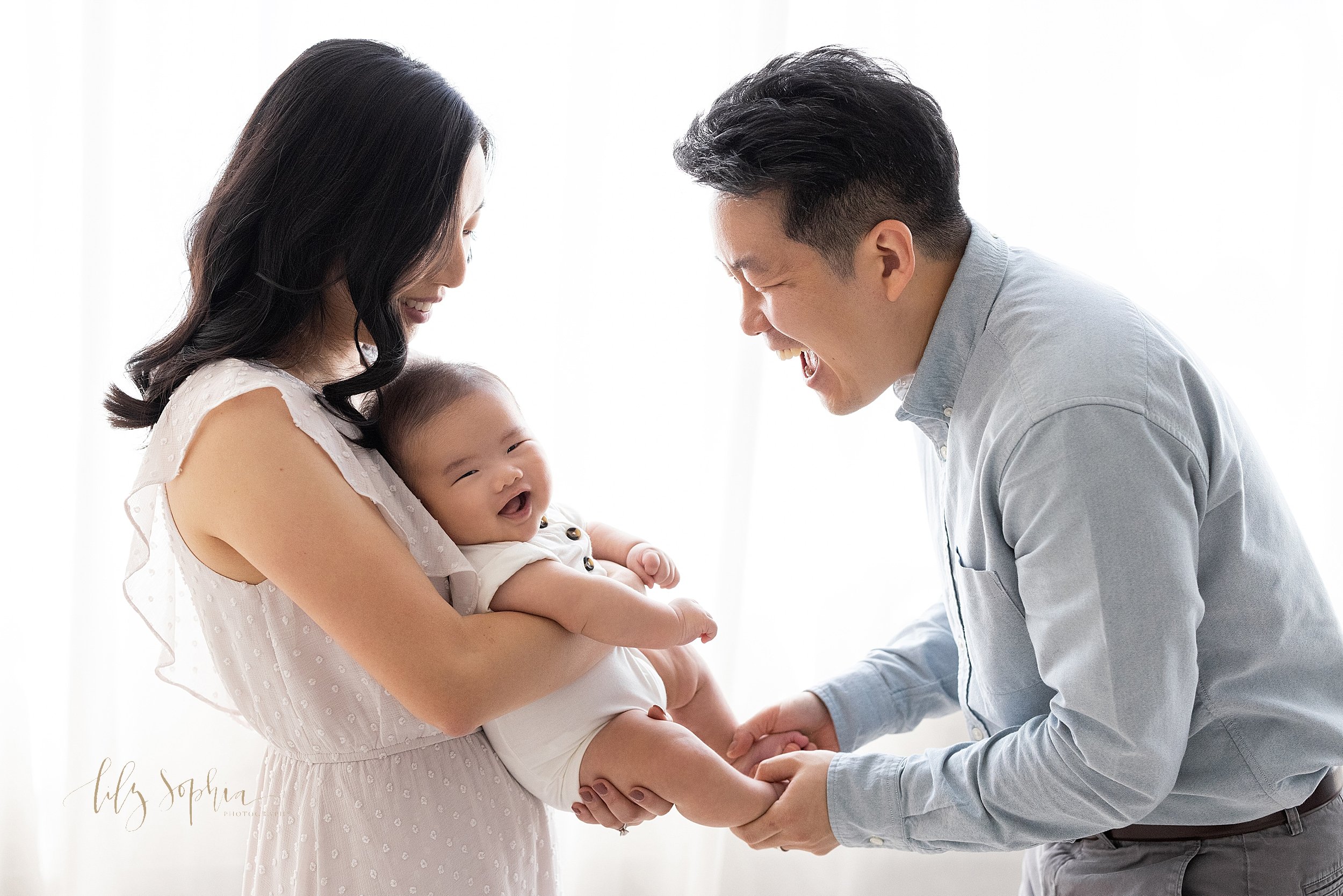  Family photo of a laughing Asian family as mom holds her son in front of her chest and dad tickles their son’s feet as the family stands in front of a window streaming natural light in a studio near Buckhead in Atlanta. 