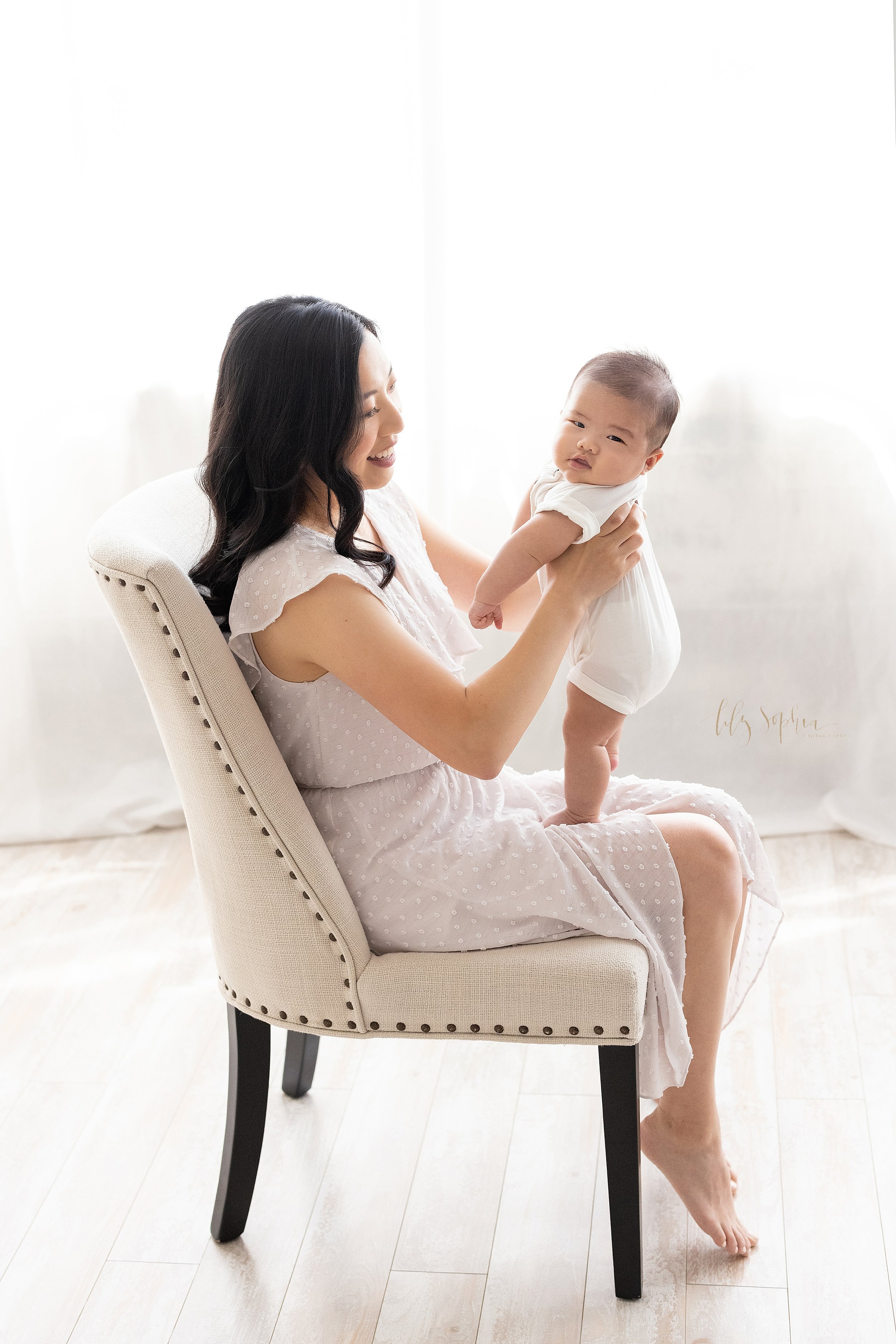  Family portrait of an Asian mother as she sits in an upholstered chair in front of a window streaming natural light and holds her infant son as he stands on her lap taken in a studio near Ansley Park in Atlanta. 