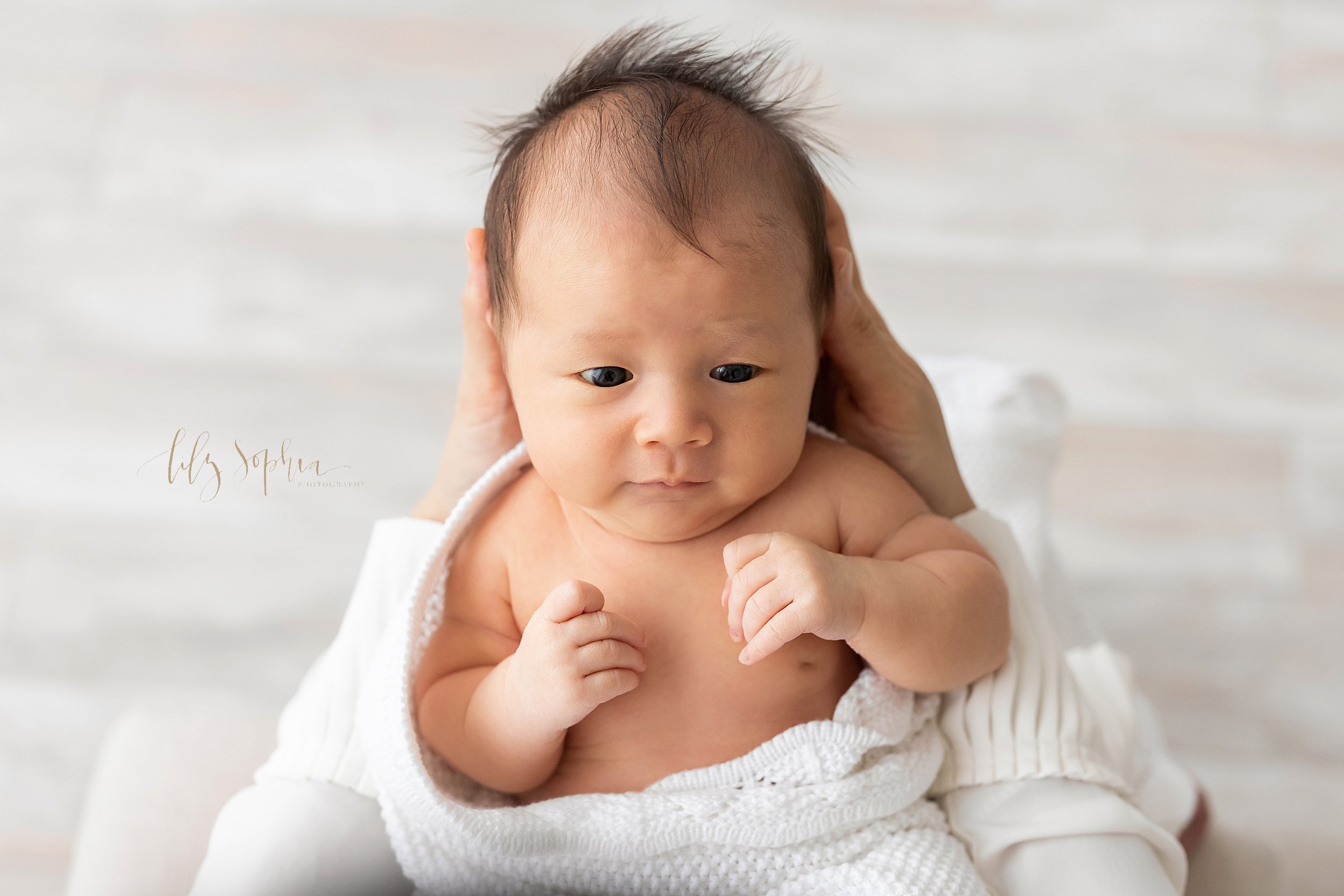  Newborn portrait of an awake infant girl with her wispy hair as she is held in her mother’s hands taken in a natural light studio near Roswell in Atlanta, Georgia. 
