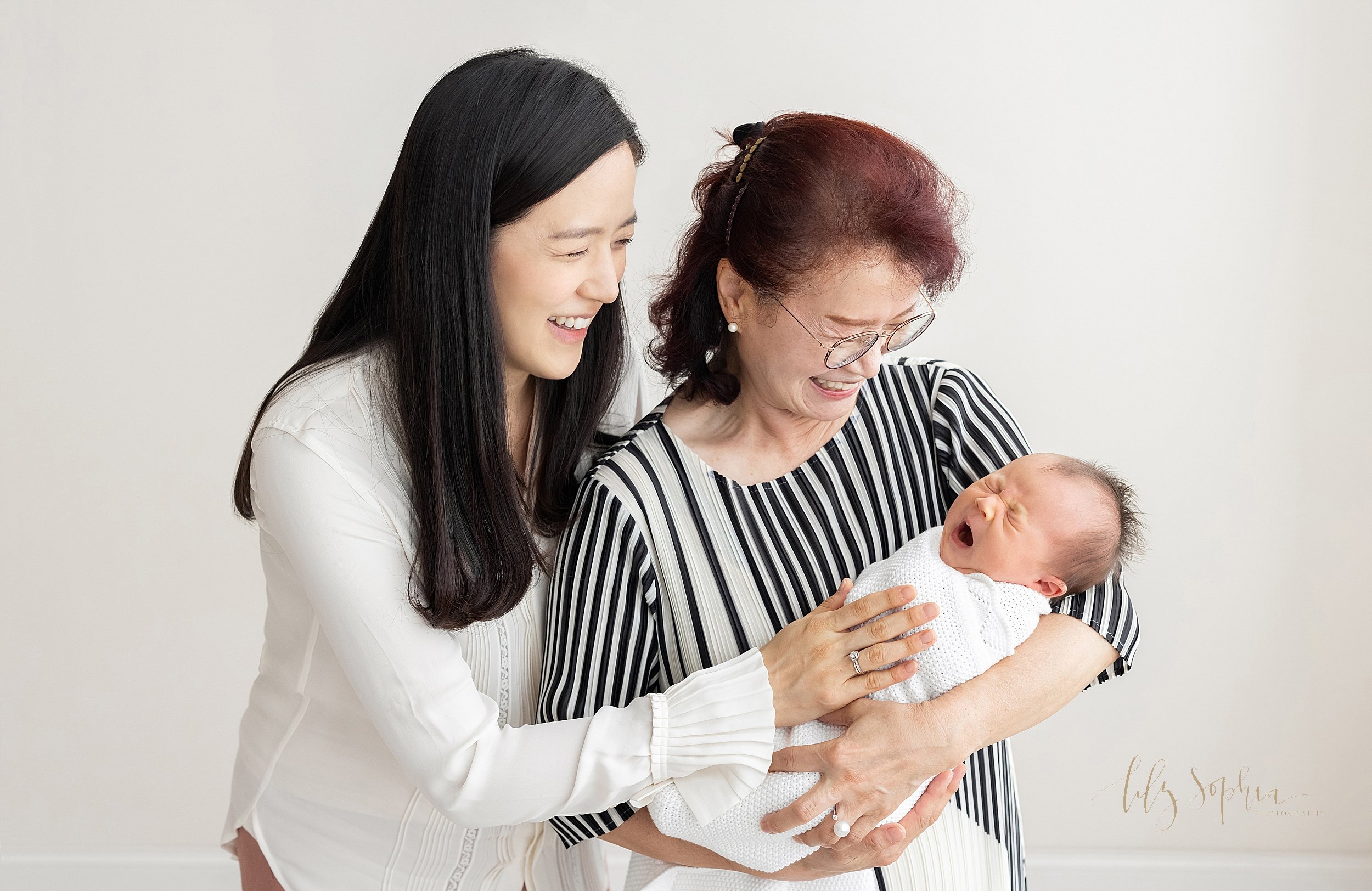  Family portrait of three generations, an Asian grandmother holding her newborn granddaughter in her arms as her daughter places her hand on her infant daughter’s chest while the newborn yawns taken in natural light in a studio near Kirkwood in Atlan