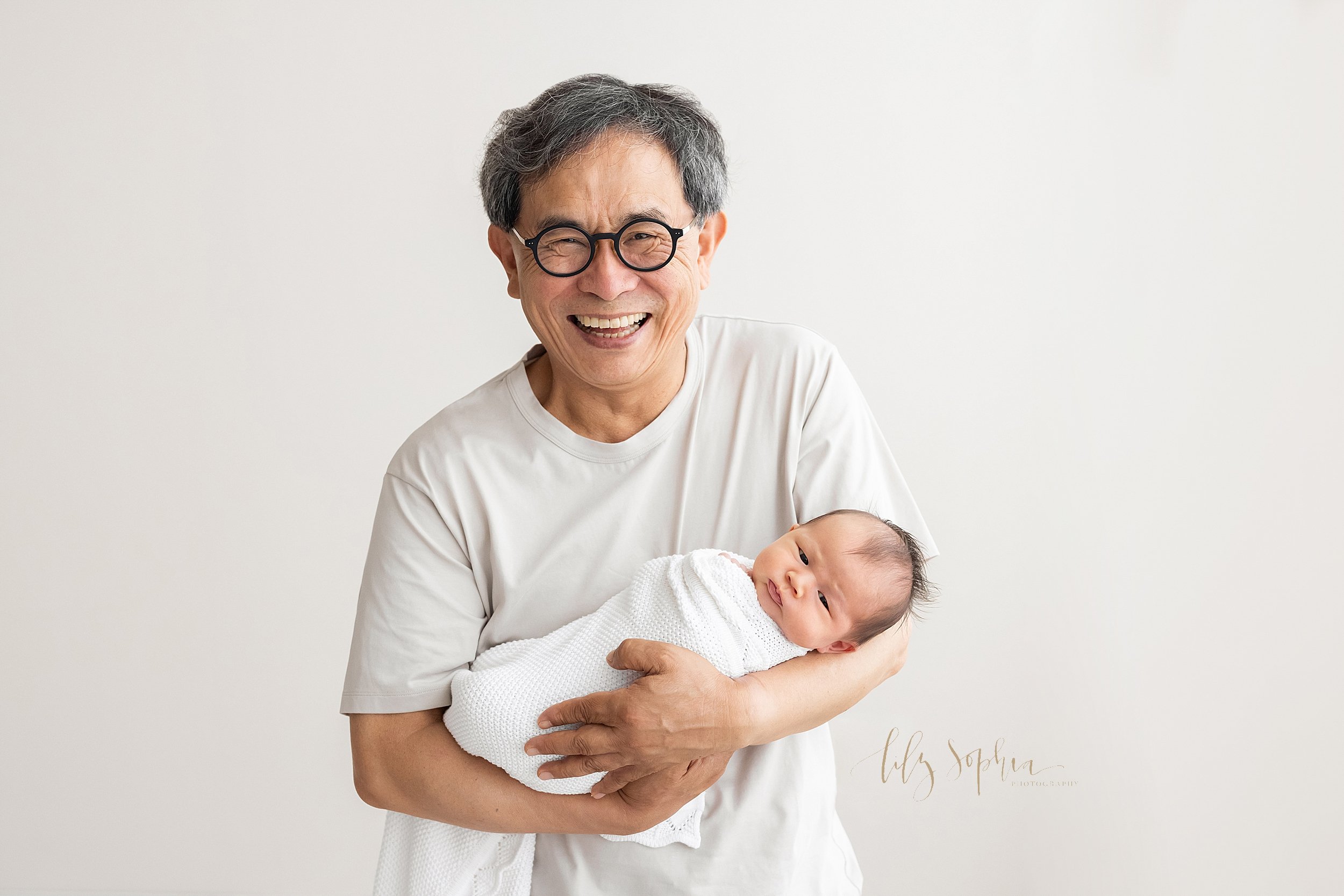  Family photo of a proud smiling Asian grandfather as he holds his newborn granddaughter in his arms taken in a natural light studio in the Ansley Park area of Atlanta. 