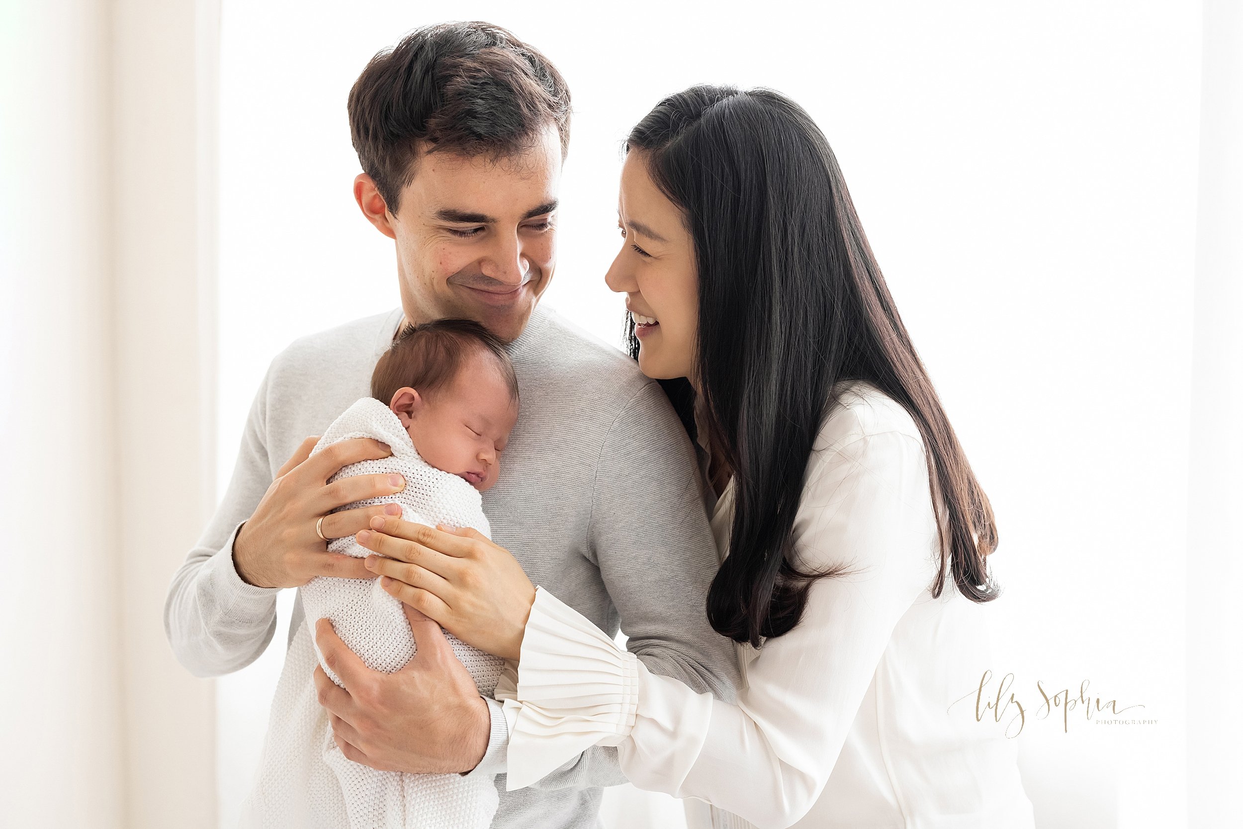  Newborn portrait of a father holding his daughter against his chest as his wife stands behind him and the parents interact with one another taken in front of a window streaming natural light in a studio near Virginia Highlands in Atlanta. 